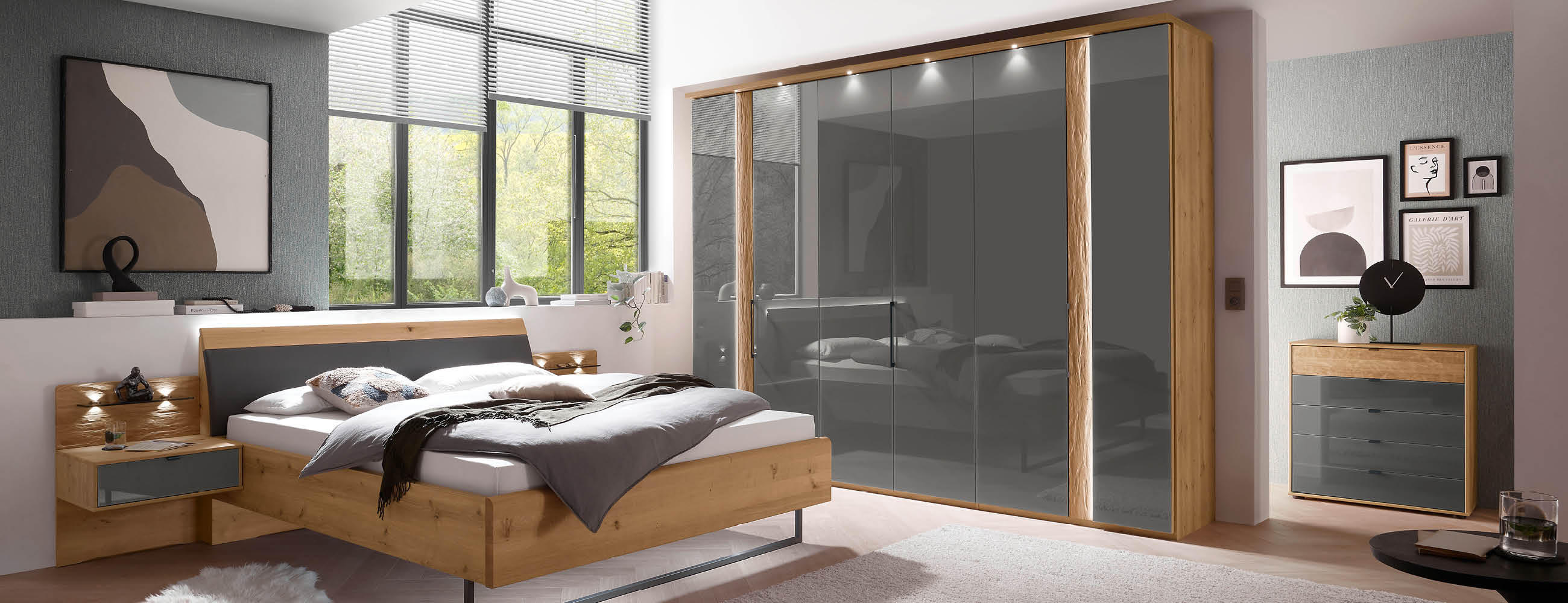 Ein Schlafzimmer mit einem Bett, einer Kommode, Kleiderschrank in der Farbe Anthrazit und einem Fenster, das Tageslicht hereinlässt.