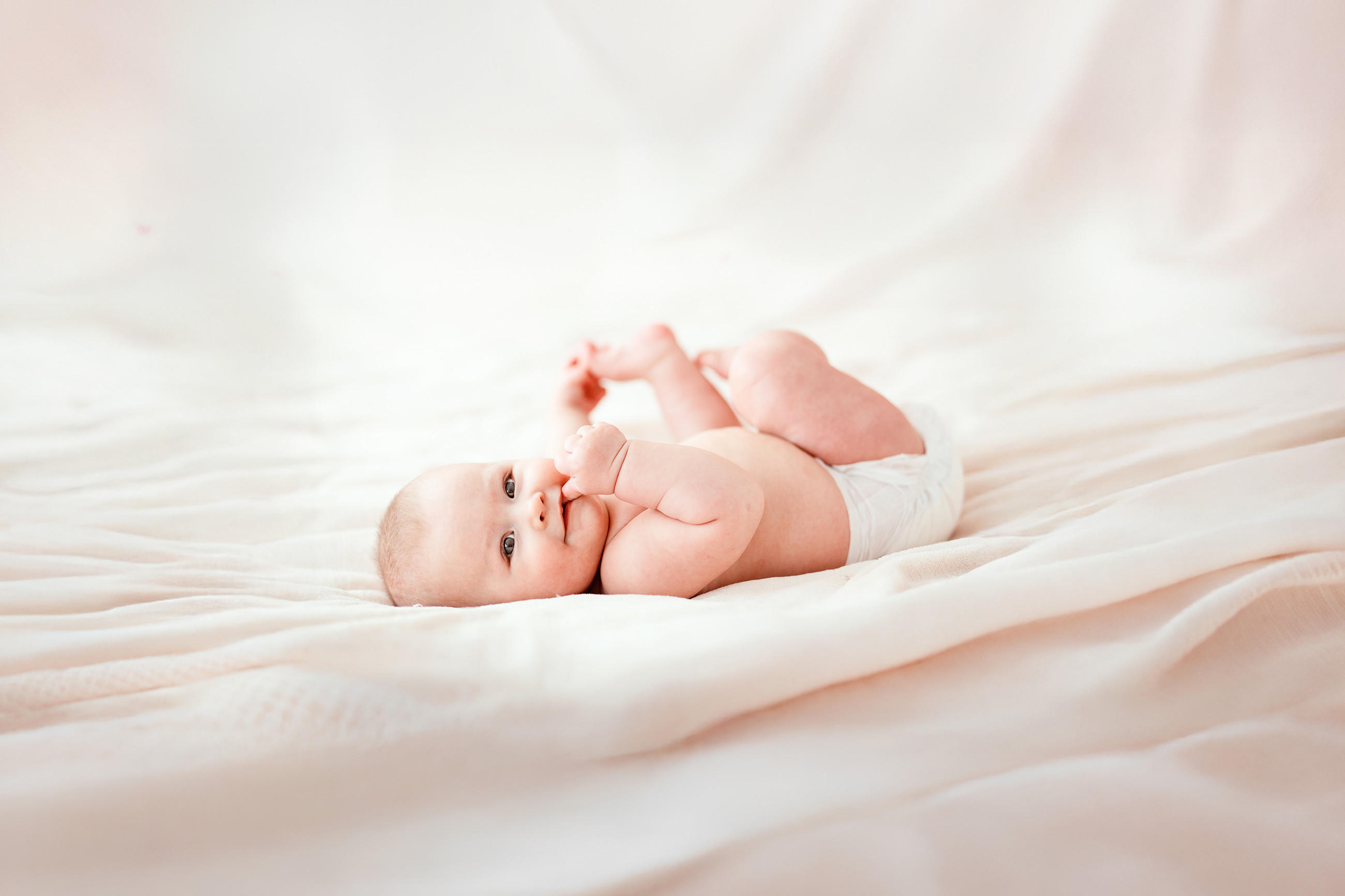 Ein Baby liegt auf dem Rücken auf weichen, cremefarbenen Decken und hebt Arme und Beine leicht an, wobei die helle Umgebung eine ruhige und sanfte Atmosphäre erzeugt.