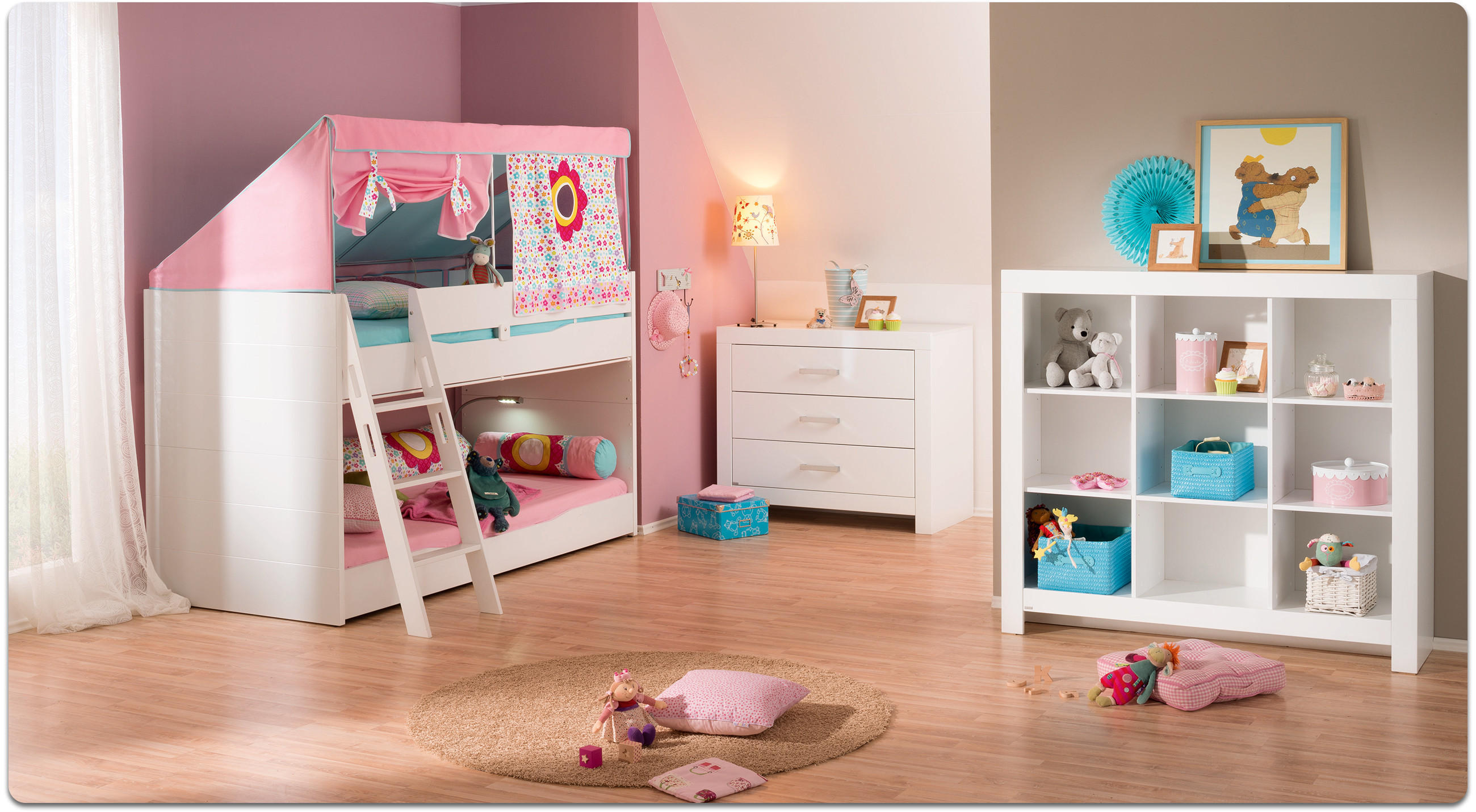 Kinderzimmer mit Etagenbett in Weiß und Rosa, Kommode, Regal mit Spielzeug und Teppich mit Kissen auf Holzboden.