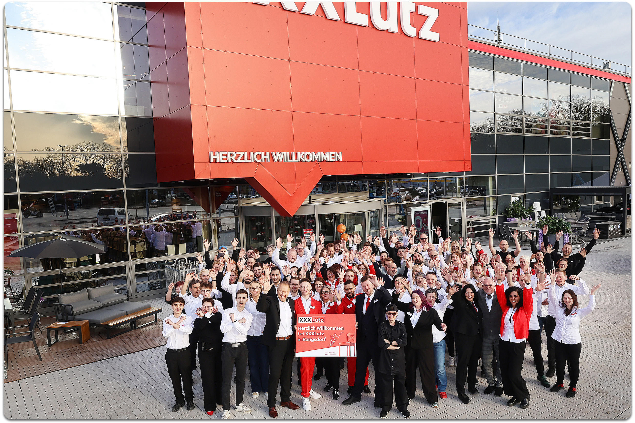 Das Team von XXXLutz Rangsdorf steht vor ihrer Filiale und alle jubeln. Einer der Mitarbeitenden hält ein Schild mit der Aufschrift: Herzlich Willkommen bei XXXLutz in Rangsdorf.