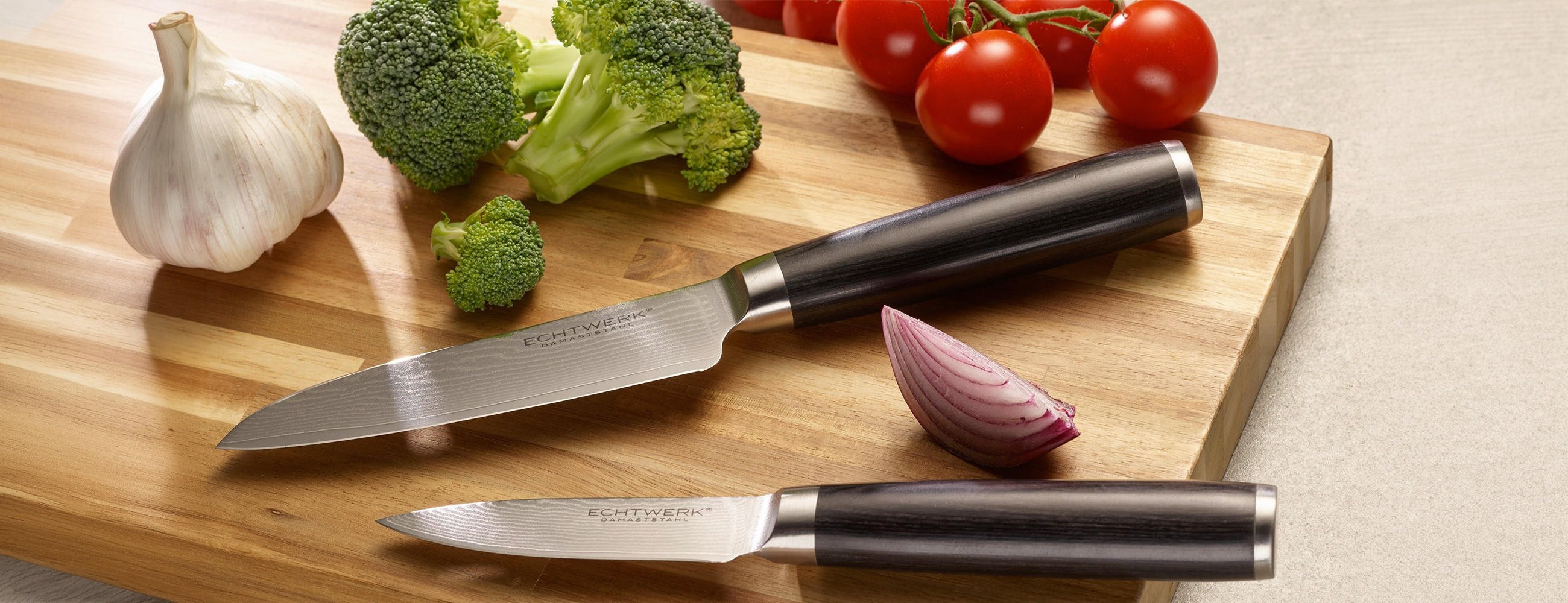 Zwei Küchenmesser mit schwarzen Griffen liegen auf einem Holzbrett neben Knoblauch, Brokkoli und Tomaten an Stielen.