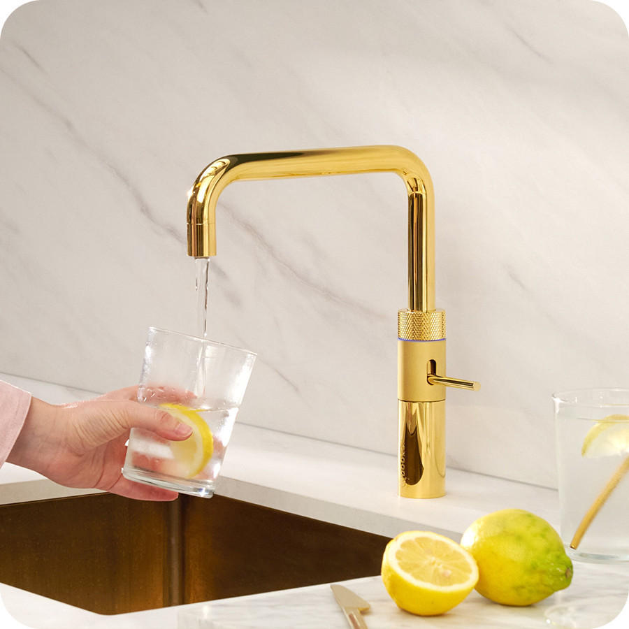 QUOOKER Küchenarmatur „Fusion Square“ in goldfarbener Ausführung mit eckigem Auslauf über einer Spüle; Wasser läuft in ein gehaltenes Glas mit Zitronenscheibe, daneben liegen Zitrone und Limette auf der Arbeitsfläche.