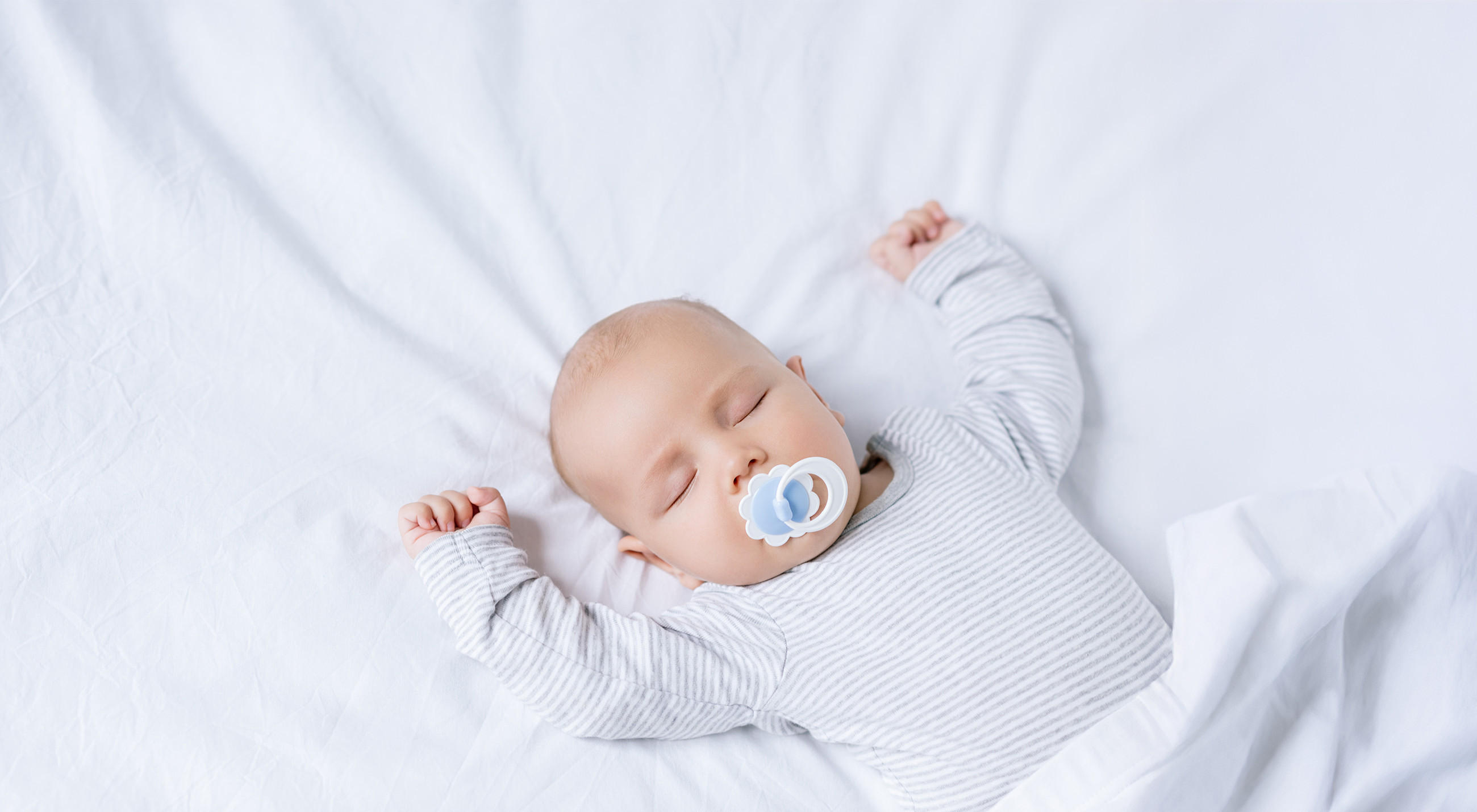 Ein Baby liegt auf dem Rücken auf weißer Bettwäsche. Die Arme sind seitlich ausgestreckt, das Baby trägt einen langärmligen, hellgrau-weiß gestreiften Einteiler. Der Kopf ist teilweise von einer Decke bedeckt.