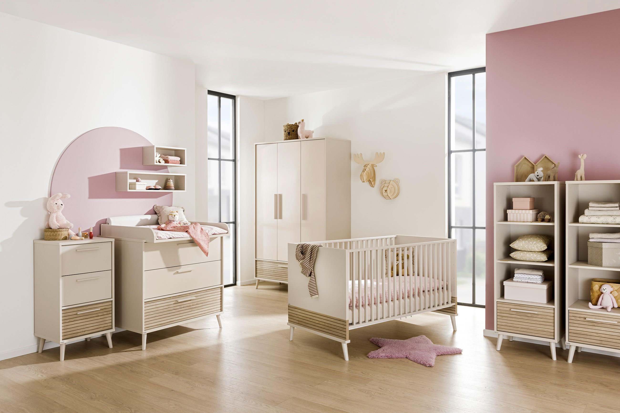 Helles Babyzimmer mit weißen Möbeln, darunter Gitterbett, Schrank und Regale. Dekoriert mit rosa Akzenten und Kuscheltieren