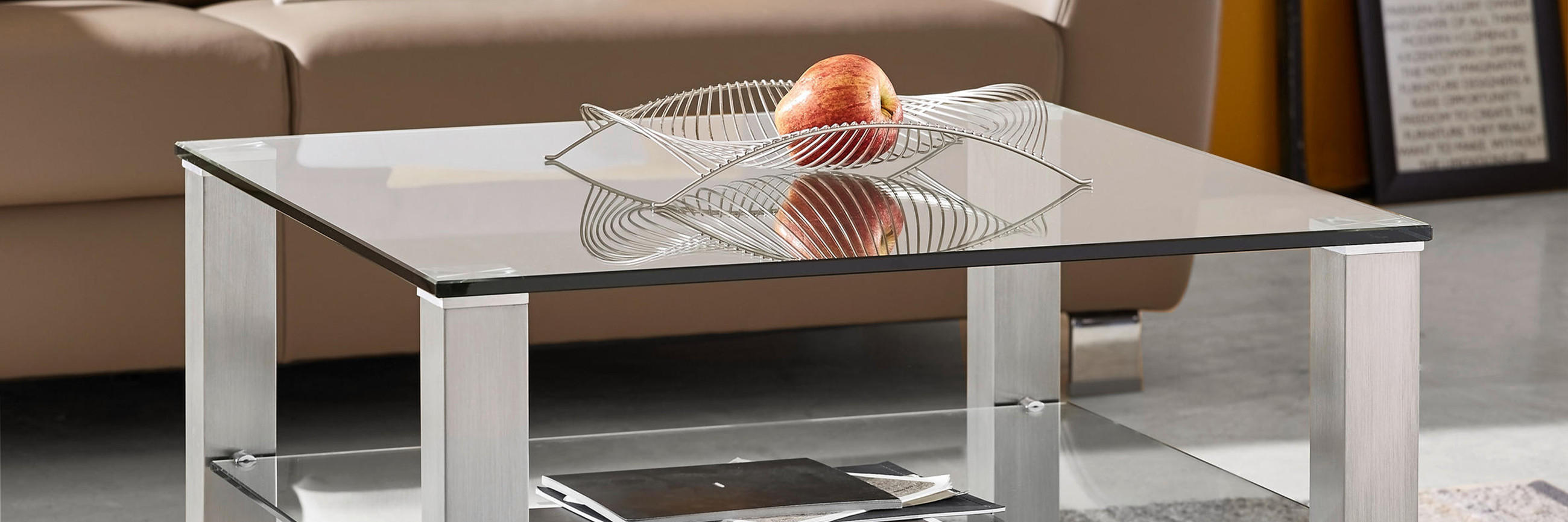 Quadratischer Couchtisch mit Glasplatte und Unterboden aus Glas, silbernen Metallbeinen und dekorativer Drahtschale mit Apfel steht auf gemustertem Teppich vor braunem Sofa