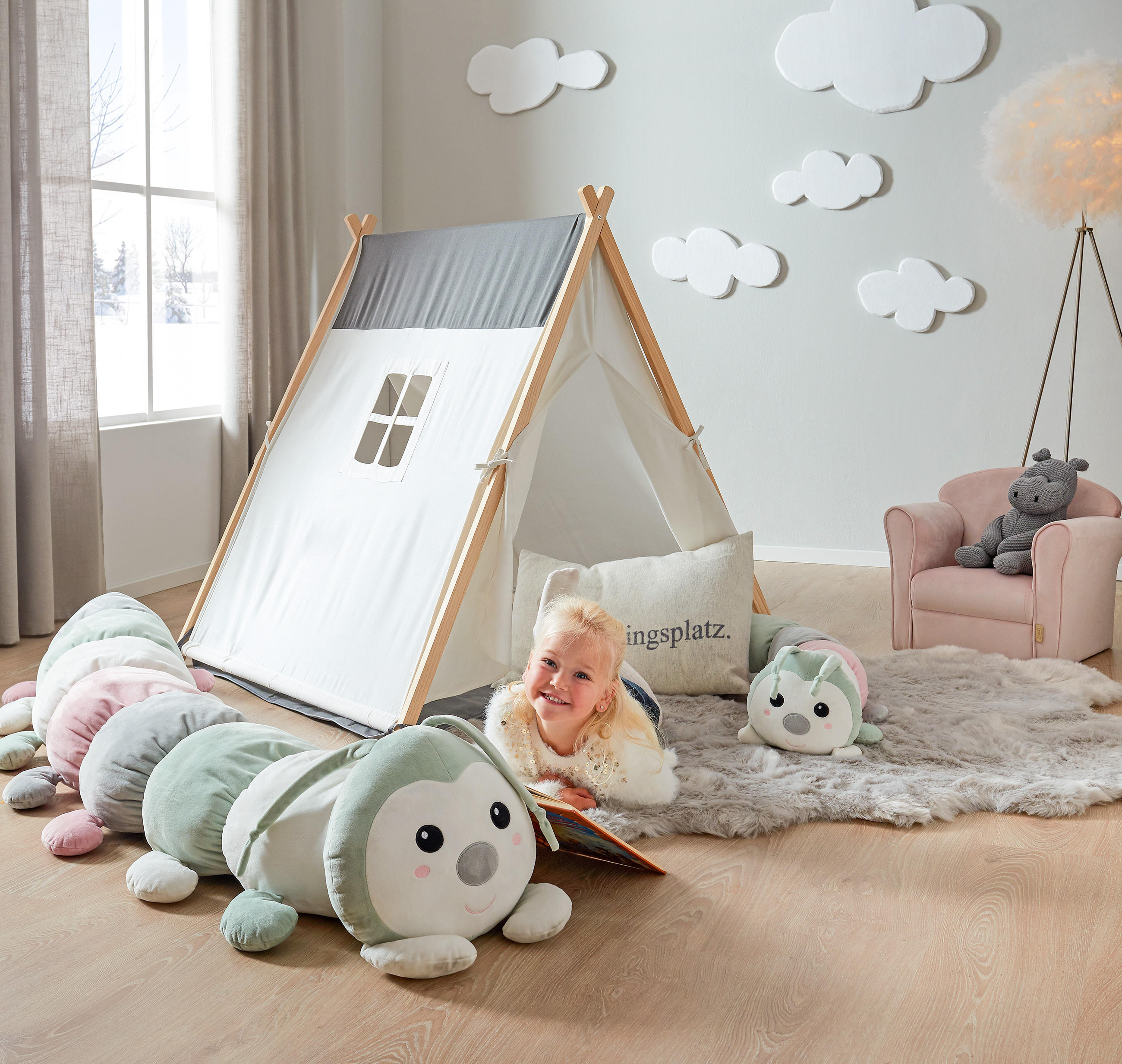 Kind liegt auf dem Boden vor einem Zeltspielhaus mit Fenster und liest ein Buch neben einem großen, weichen Raupenkuscheltier in einem hellen Kinderzimmer mit Teppich, Sessel und Wolkenwanddekoration.