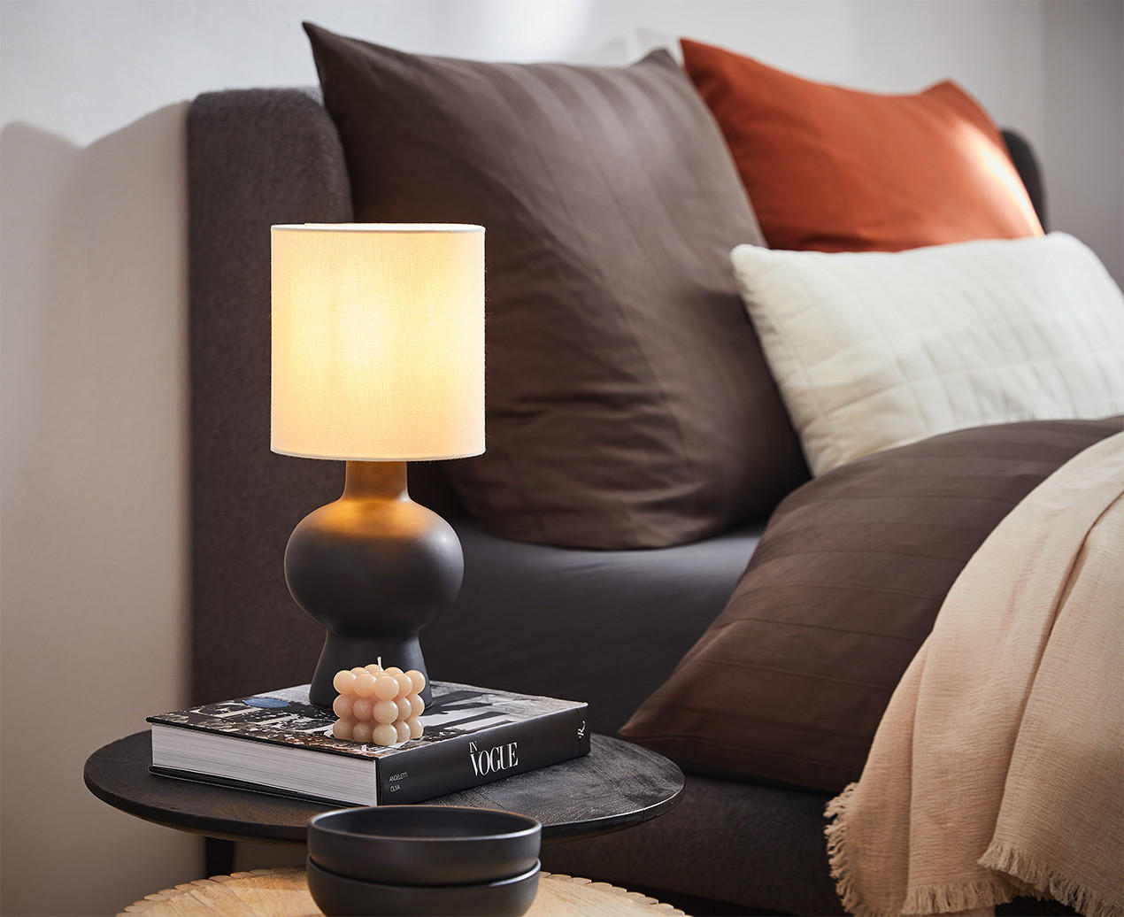 Gemütliches Schlafzimmer mit Nachttisch, Lampe, Deko-Kerze und Kissen in warmen Farben