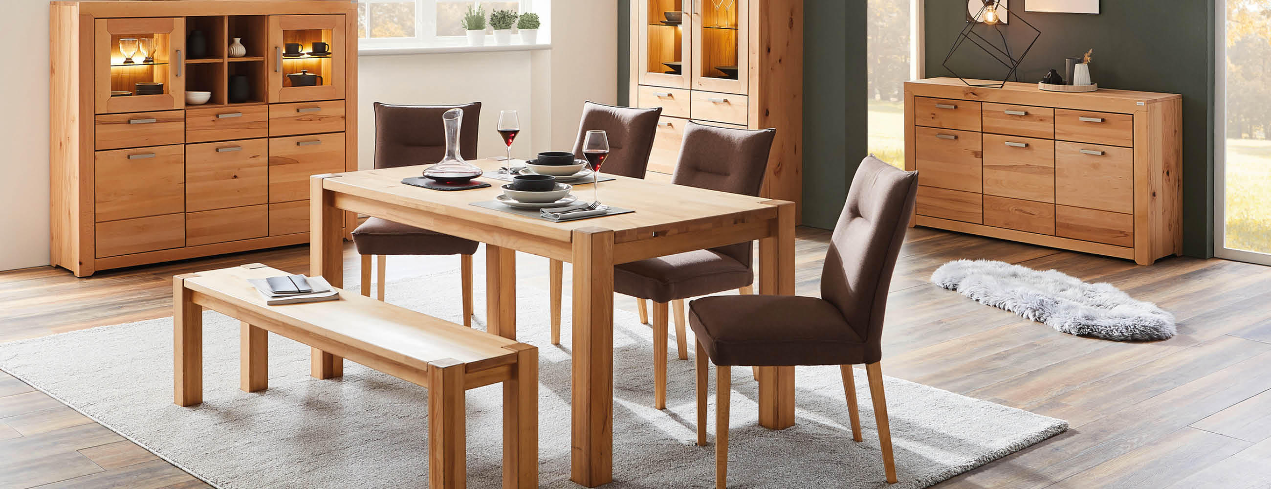 Esszimmer mit hellem Holztisch, zwei gepolsterten Stühlen, einer Holzbank, zwei Vitrinen und einem Sideboard auf hellem Teppichboden