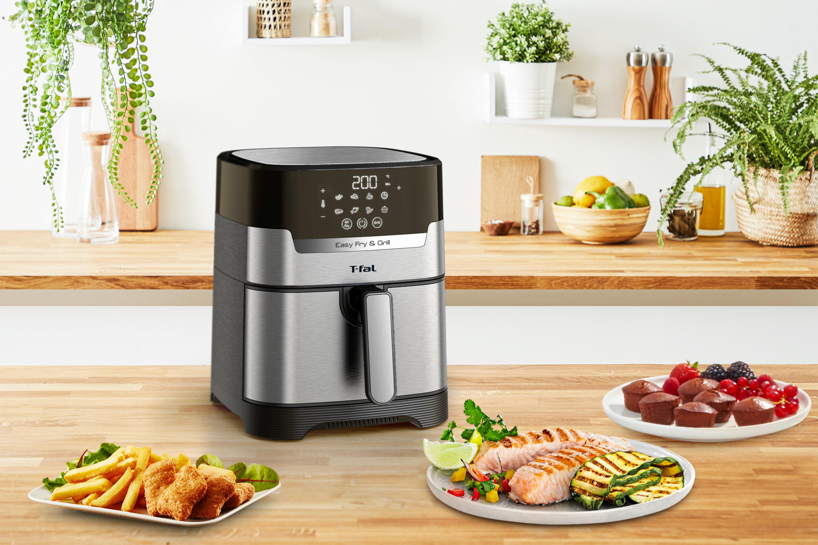 Eine Heißluftfriteusse von TEFAL steht auf einer Arbeitsplatte aus Holz, zusammen mit 3 Tellern die unterschiedliche Gerichte zeigen.