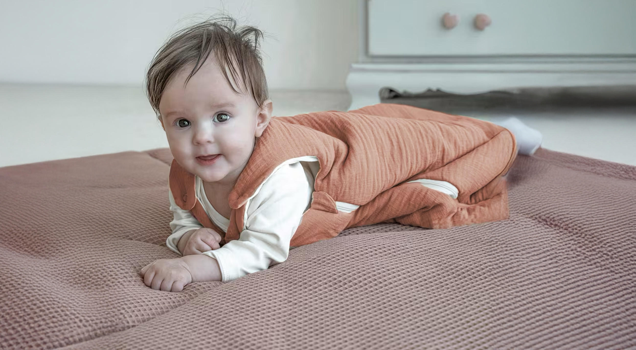 Ein Baby liegt auf einer weichen Spielmatte und trägt einen orangefarbenen Schlafsack über weißer Kleidung; im Hintergrund ist ein Möbelstück mit zwei Knöpfen zu sehen