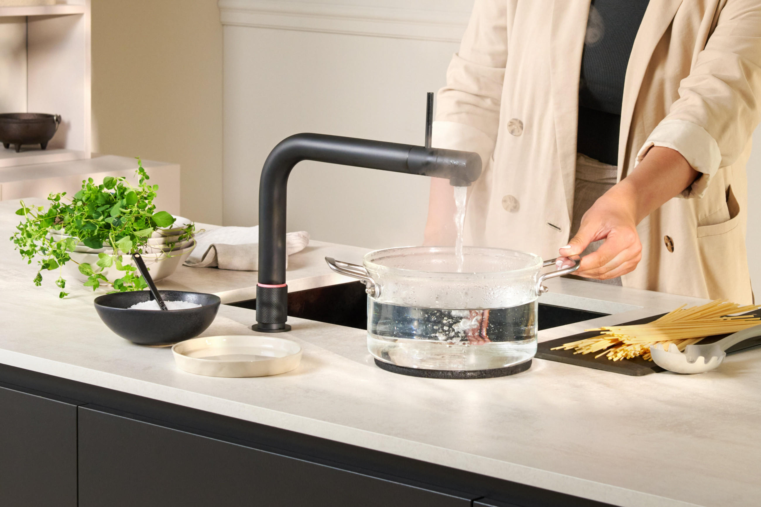 Quooker Wassersystem-Armatur in mattem Schwarz füllt einen Topf mit heißem Wasser am Spülbecken; daneben liegen frische Kräuter, Salz und Pasta auf der Arbeitsfläche.