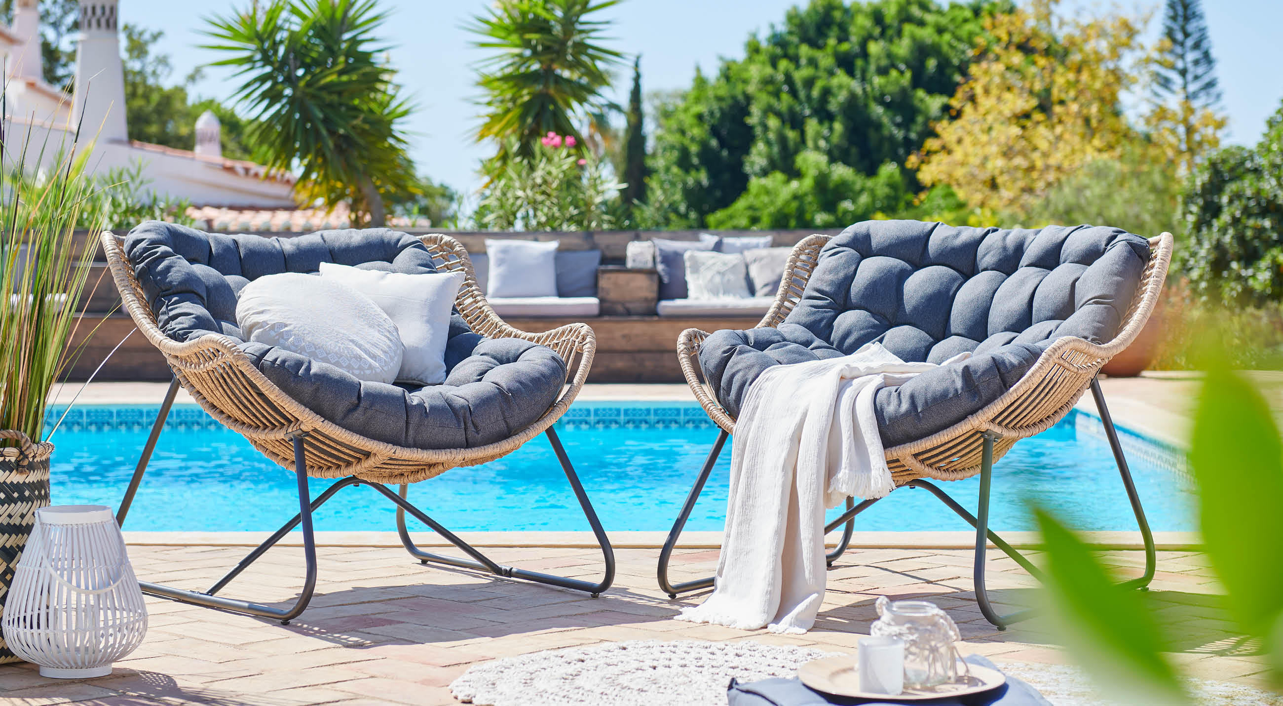 Zwei runde Loungesessel mit grauen Polstern und Rattanrahmen am Pool, im Hintergrund Palmen und Sitzbank.