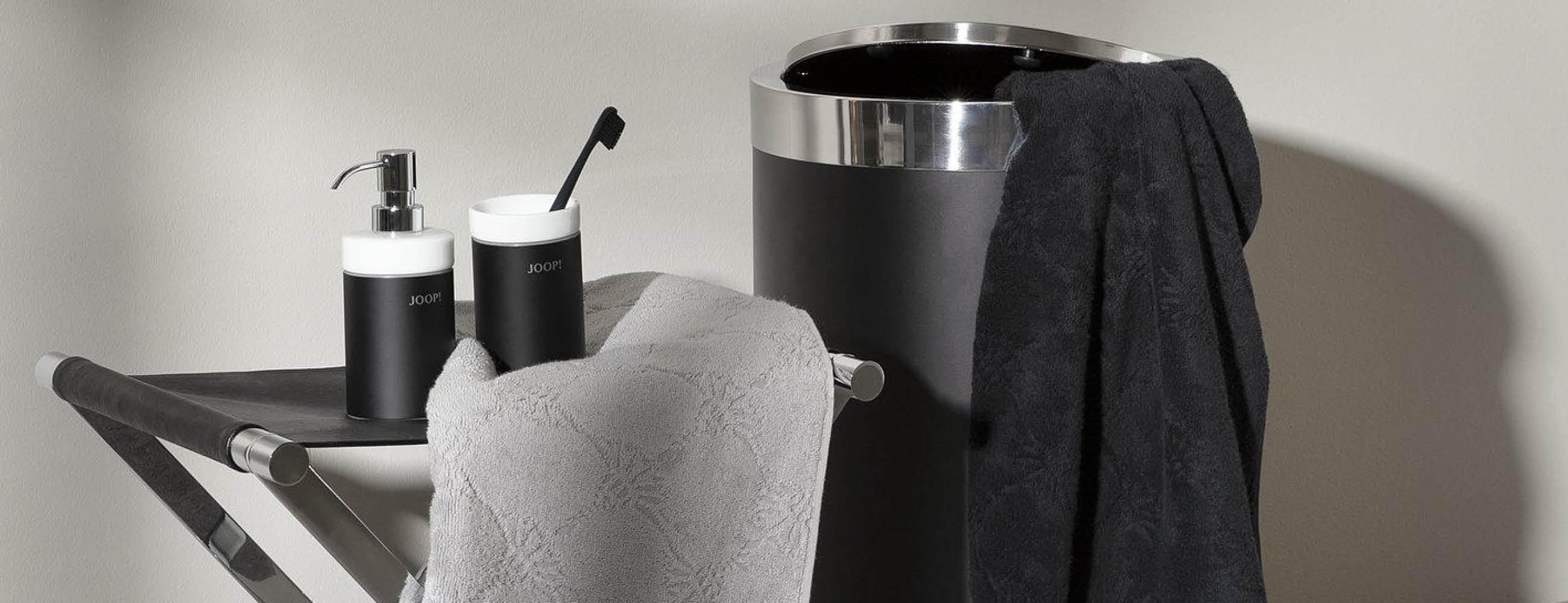 Badezimmer mit schwarzem Handtuchhalter, grauem Handtuch, schwarzem Seifenspender und schwarzer Zahnbürste auf Ablage sowie schwarzem Wäschekorb