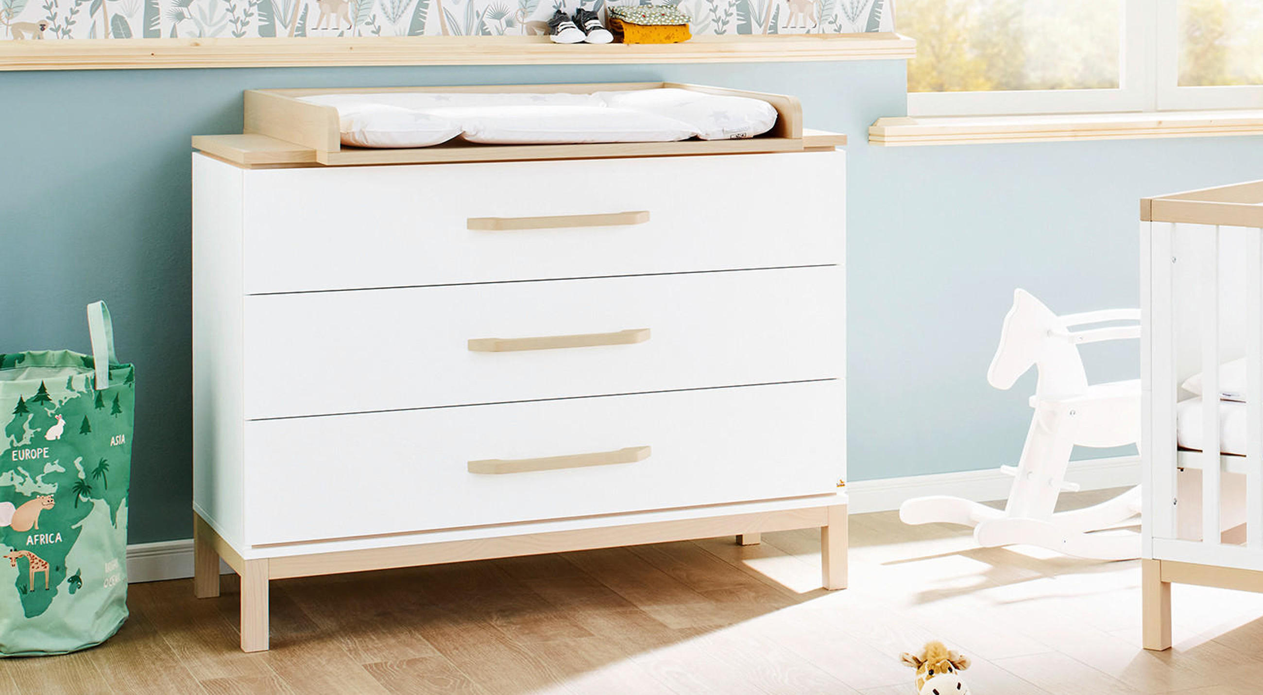 Weiße Wickelkommode mit Wickelauflage in einem hellblau gestrichenen Kinderzimmer, daneben ein grüner Aufbewahrungssack mit Tiermotiven und ein weißes Schaukelpferd