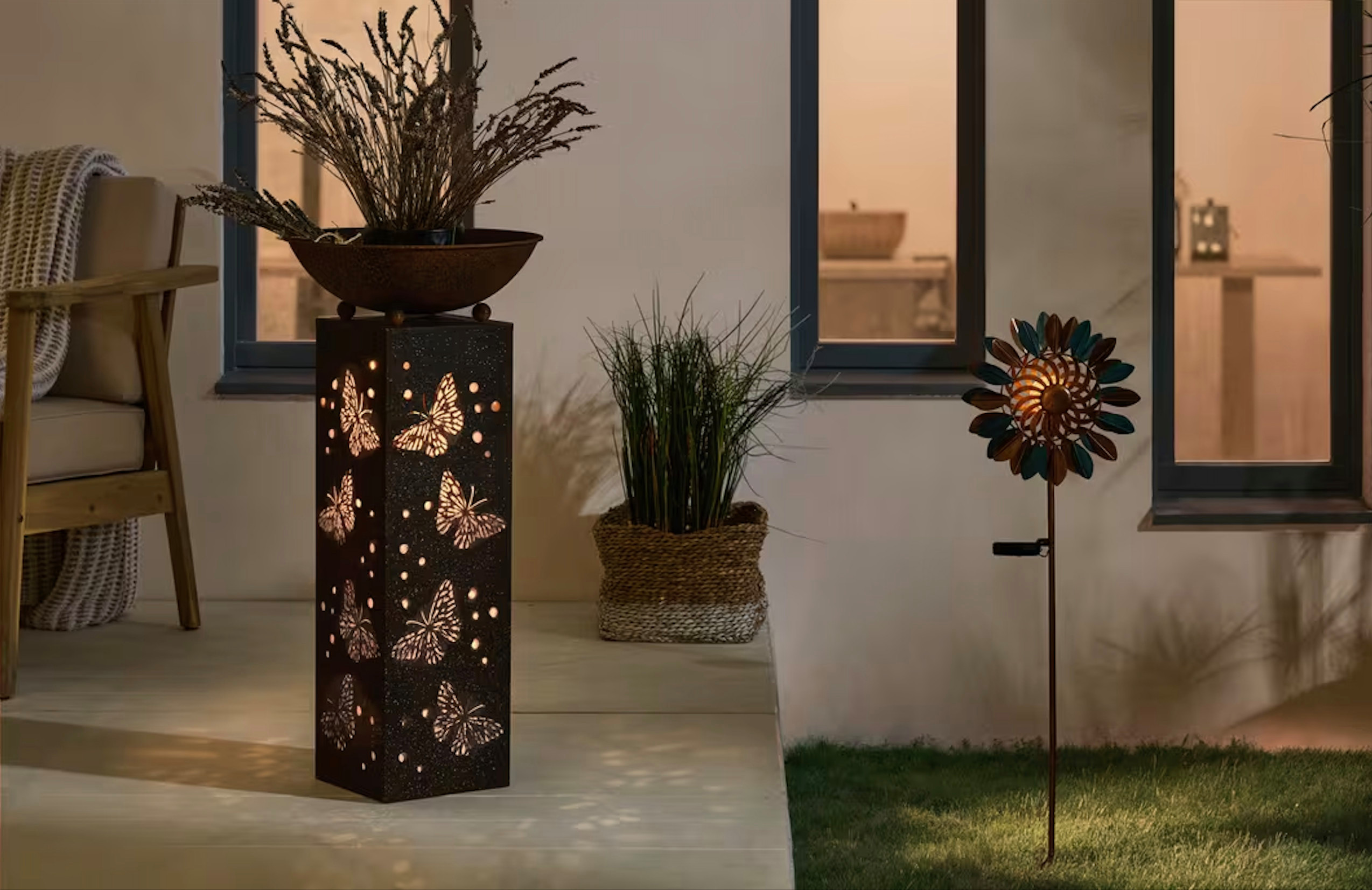 Zwei Solarleuchten und eine Feuerschale in Rostoptik auf einer Terrasse in einem Garten im Hintergrund ein brauner geflochtener Korb als Blumentopf für grünes Dekogras