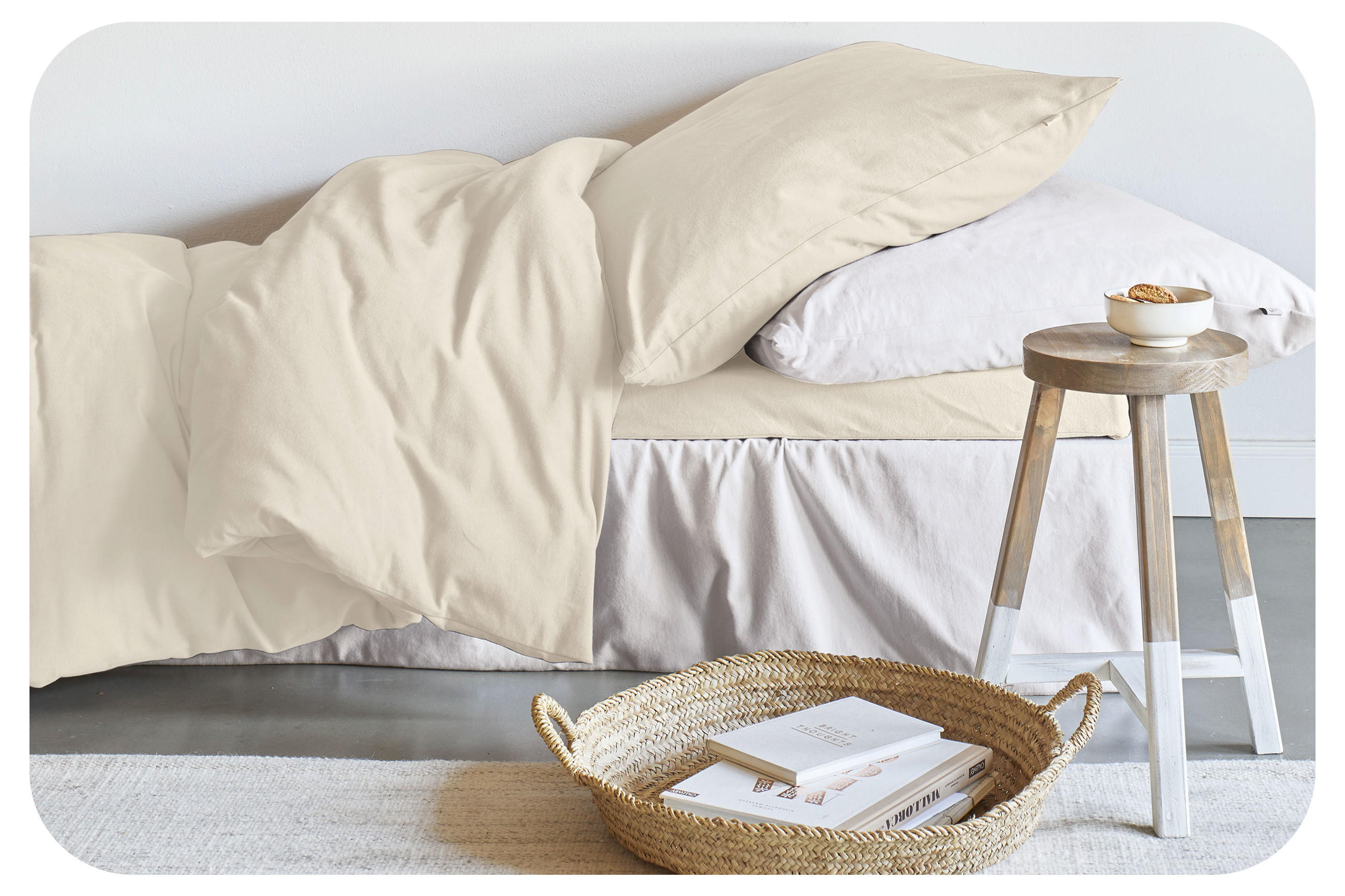 Útulná posteľ s bielou a béžovou bielizňou a vankúšom, pred ktorou stojí košík a stolička, ktoré slúžia ako praktický úložný priestor.