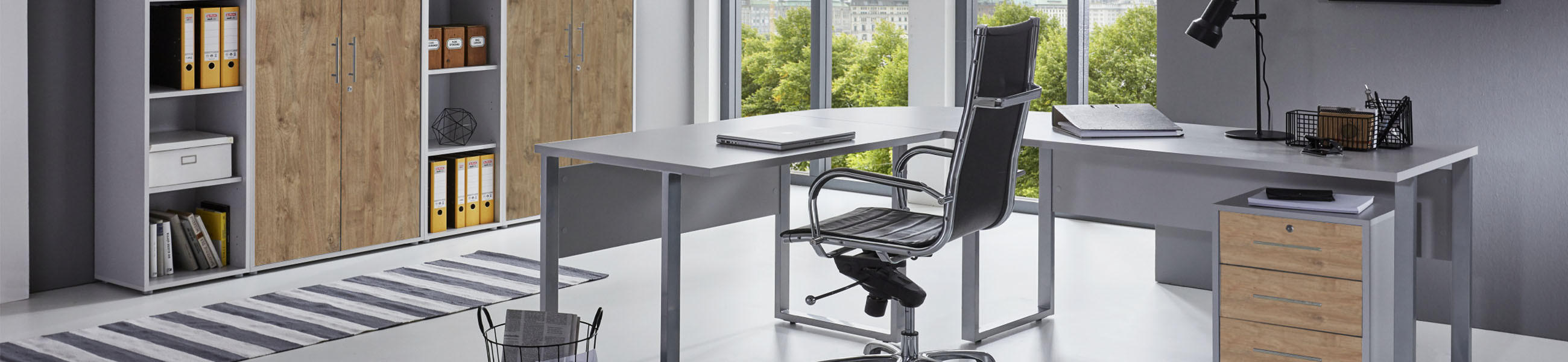 Modernes Büro mit Eckschreibtisch, schwarzem Bürostuhl, Rollcontainer und braunen Aktenschränken vor großen Fenstern.