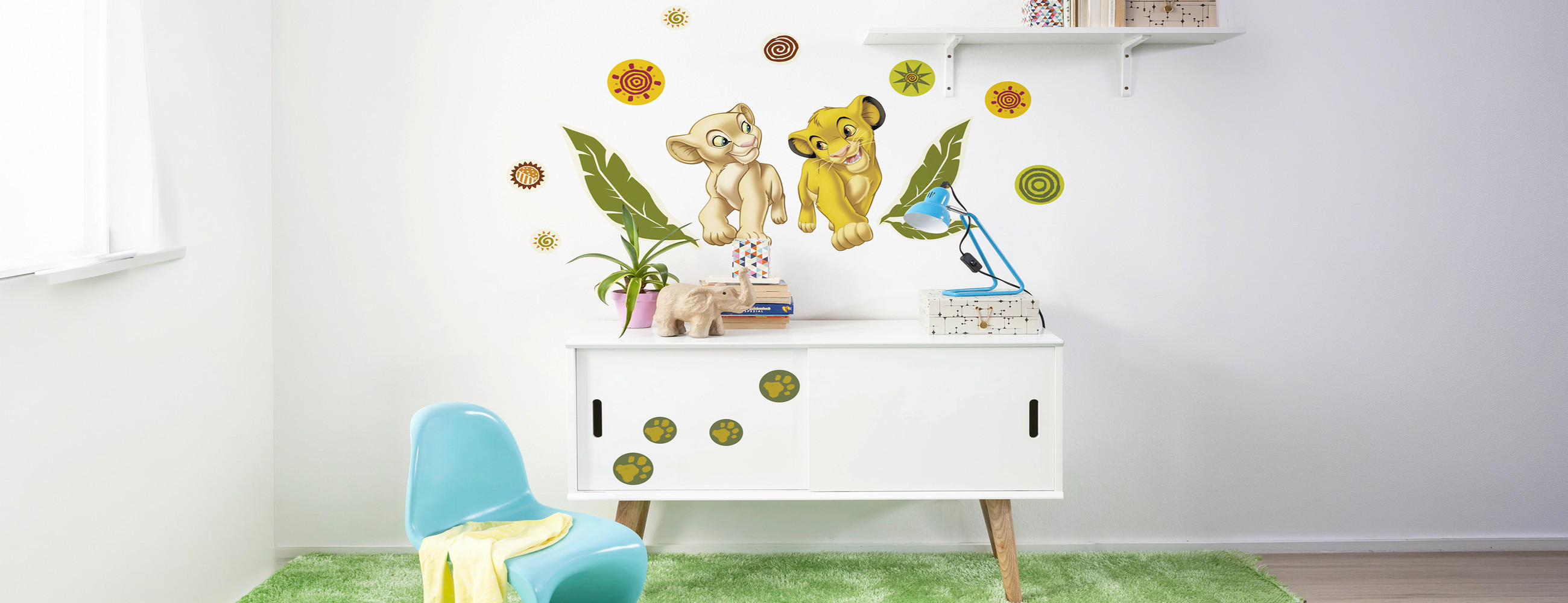 Kinderzimmer mit grünem Teppich, weißem Sideboard, blauem Stuhl, Wandsticker mit zwei Löwenbabys, Blättern und bunten Kreisen.