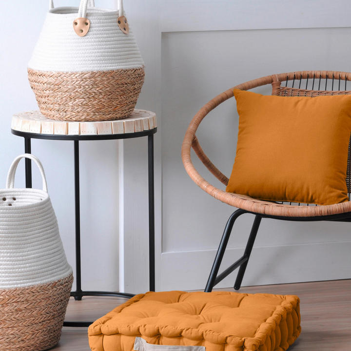 Oranžna blazina na stolu, sedežna blazina na tleh in košara iz vrbovine ob stolu.