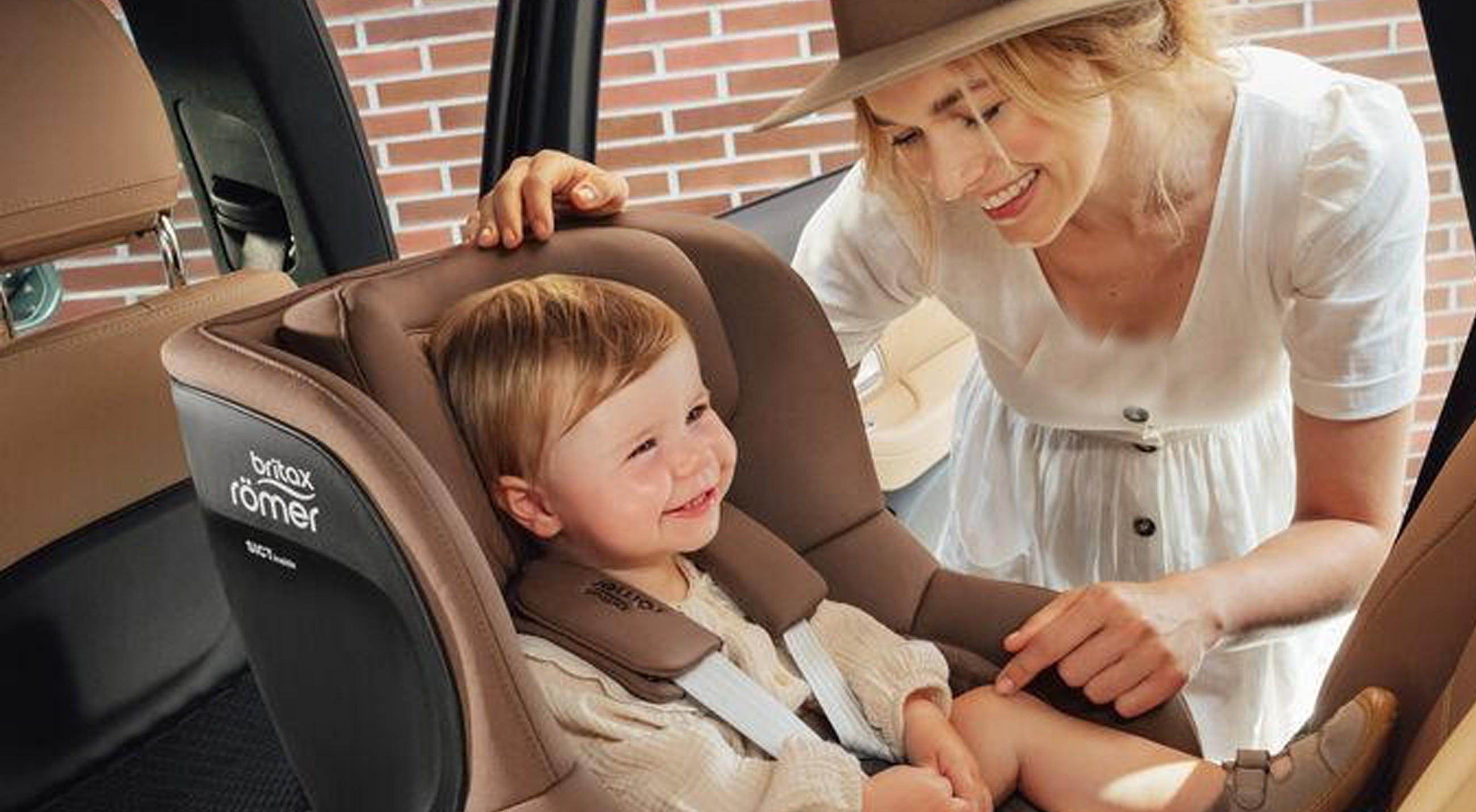 Ein Kind sitzt sicher in einem Britax Römer Kindersitz im Auto, während eine erwachsene Person sich von außen zum Kind hineinbeugt; im Hintergrund sind das Autoinnere und eine Backsteinwand zu sehen