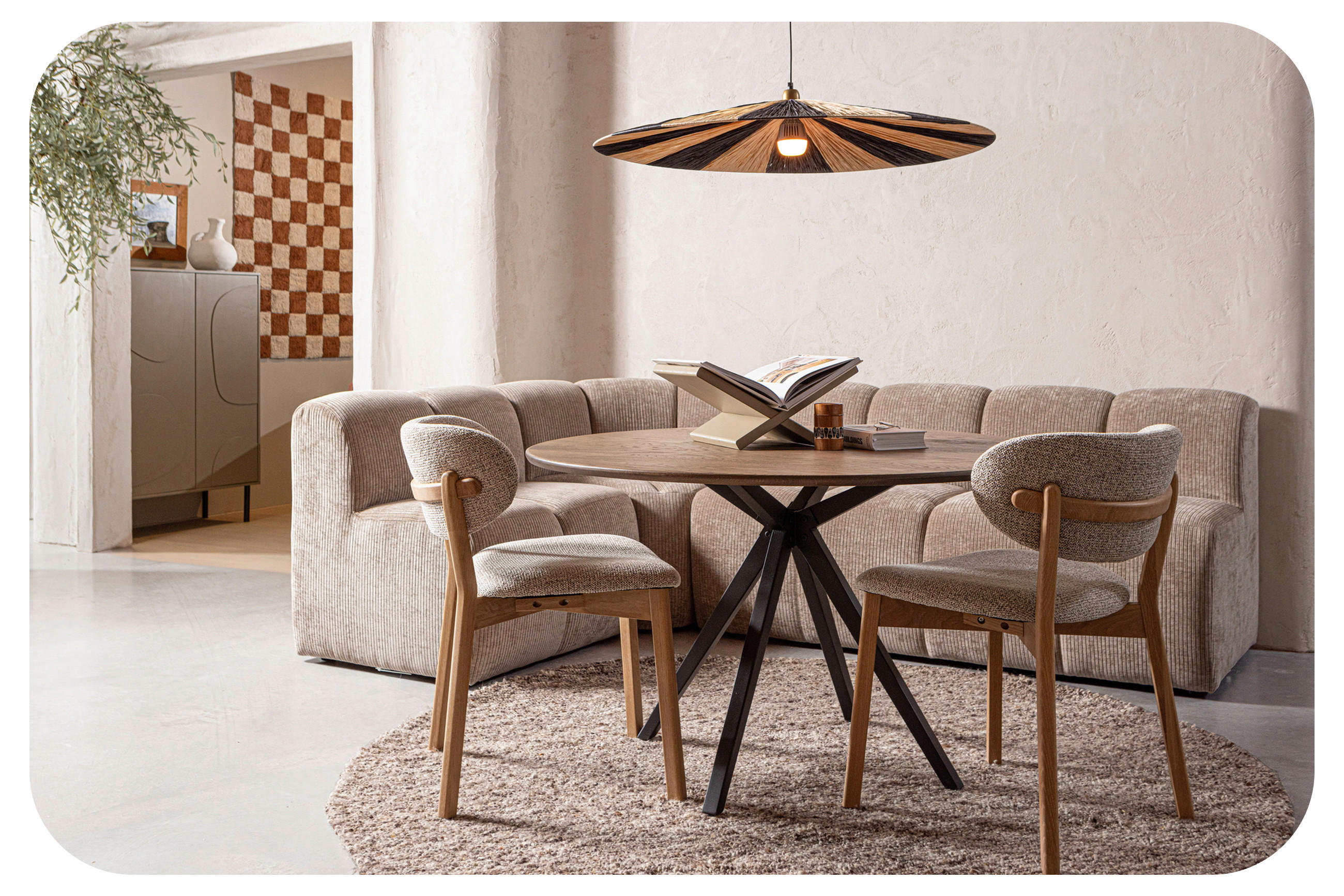 Helles Wohn- und Essmilieu mit Rundätisch, zwei Stühlen, Sofa, Teppich und großer Pendelleuchte; im Hintergrund Sideboard und Wandteppich.