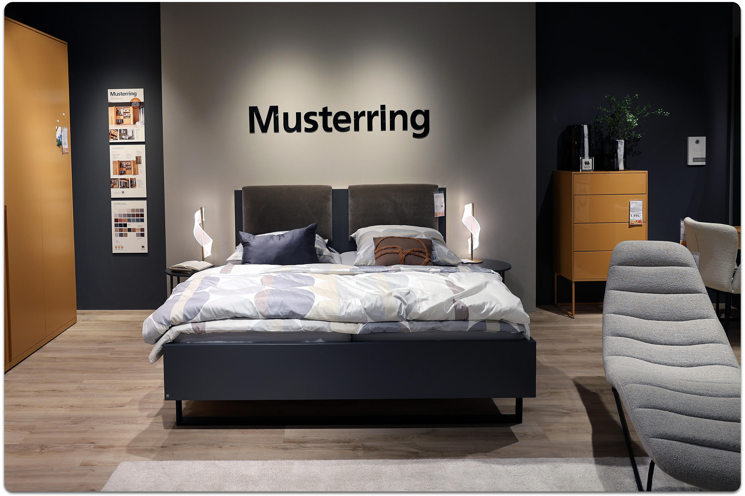 Bettenausstellung der Marke Musterring mit Markenschriftzug an der Wand in schwarz und grauem Bett im Vordergrund.