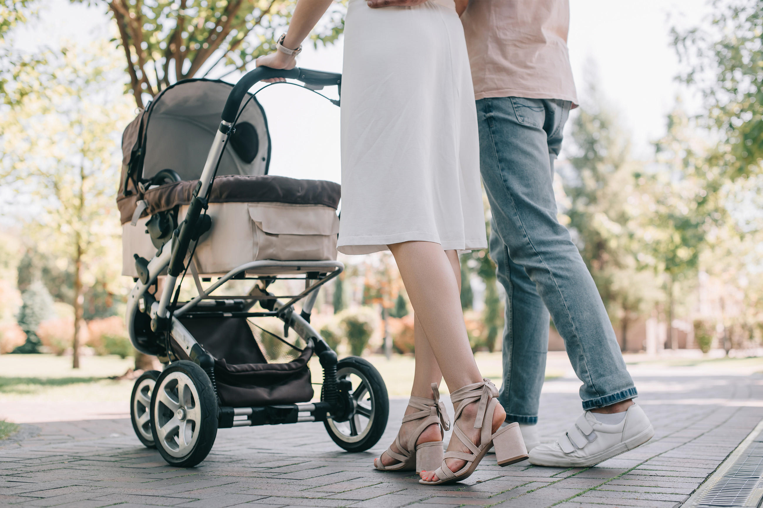 Zwei Personen stehen neben einem beige-schwarzen Kinderwagen mit großen Rädern in einem grünen Außenbereich, sichtbar nur von der Hüfte abwärts