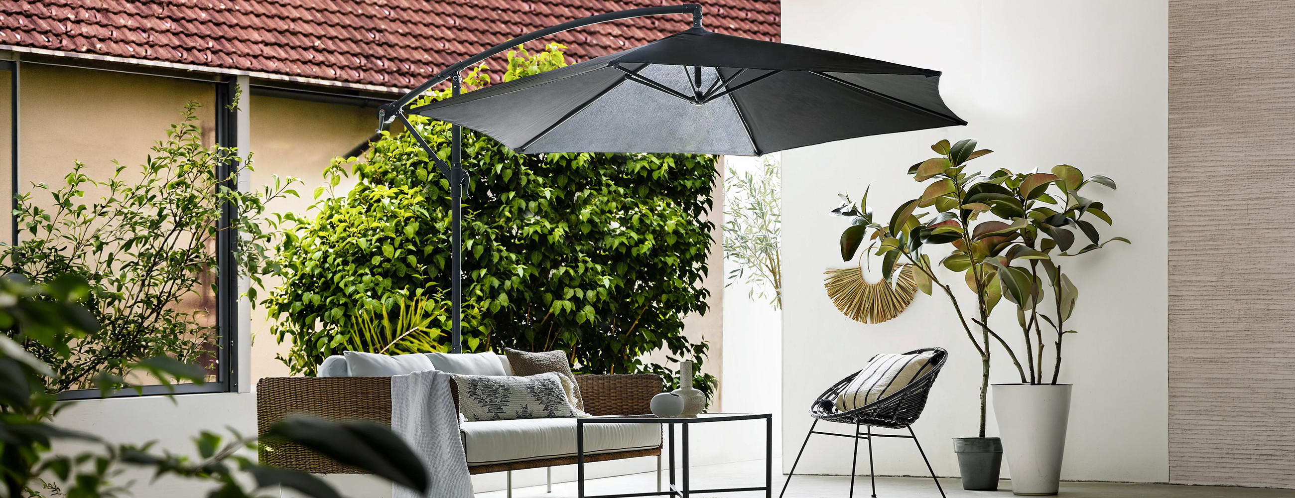 Moderner Sitzbereich im Außenbereich mit Sofa, Stuhl, großem Sonnenschirm und Pflanzen vor einer Hauswand.