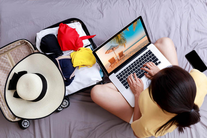Eine Frau auf einem Bett mit Laptop und Hut, die gerade ihren Urlaub plant.