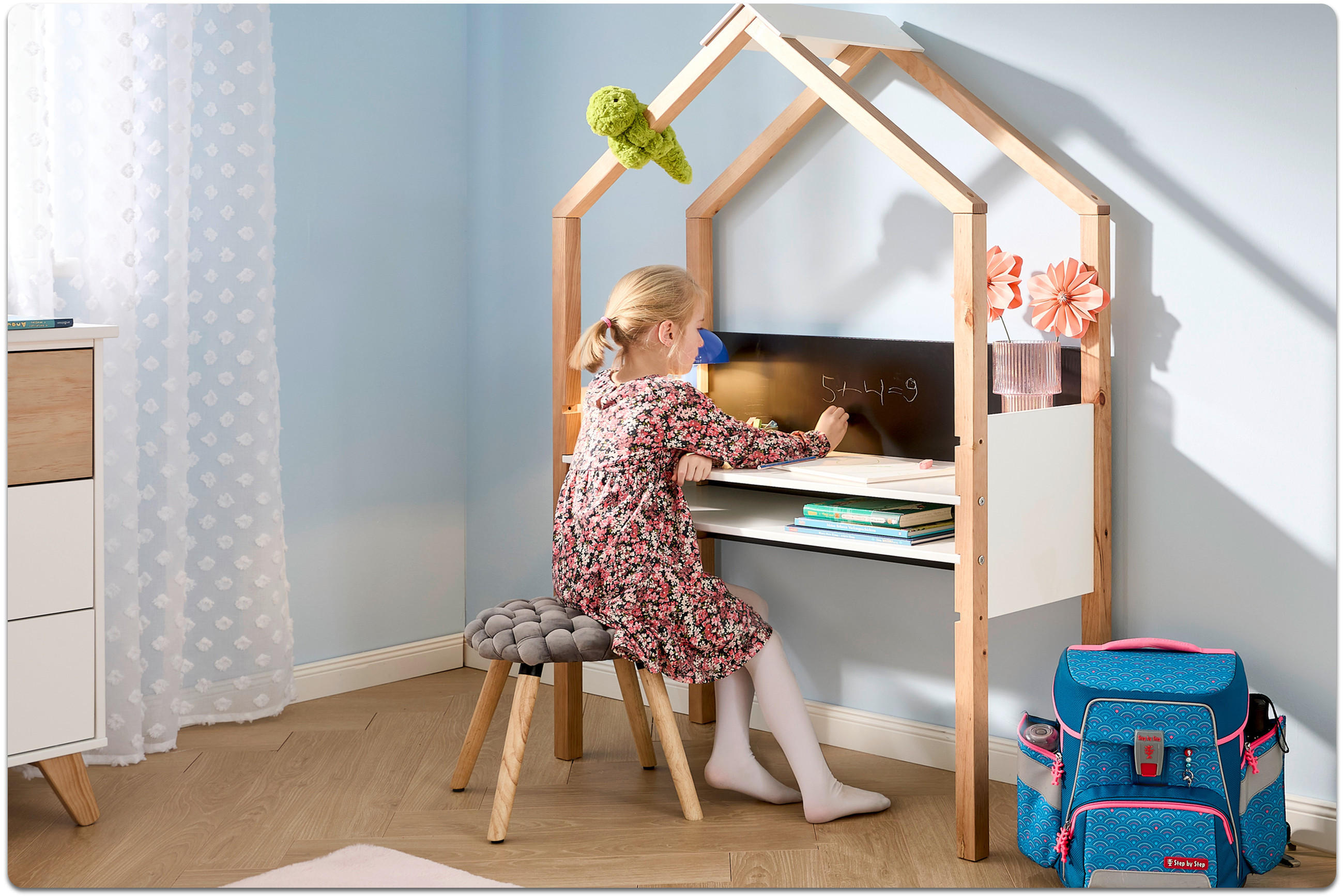 Kind sitzt auf einem gepolsterten Hocker vor einem hölzernen Kinderschreibtisch mit Tafel und schreibt mit Kreide.