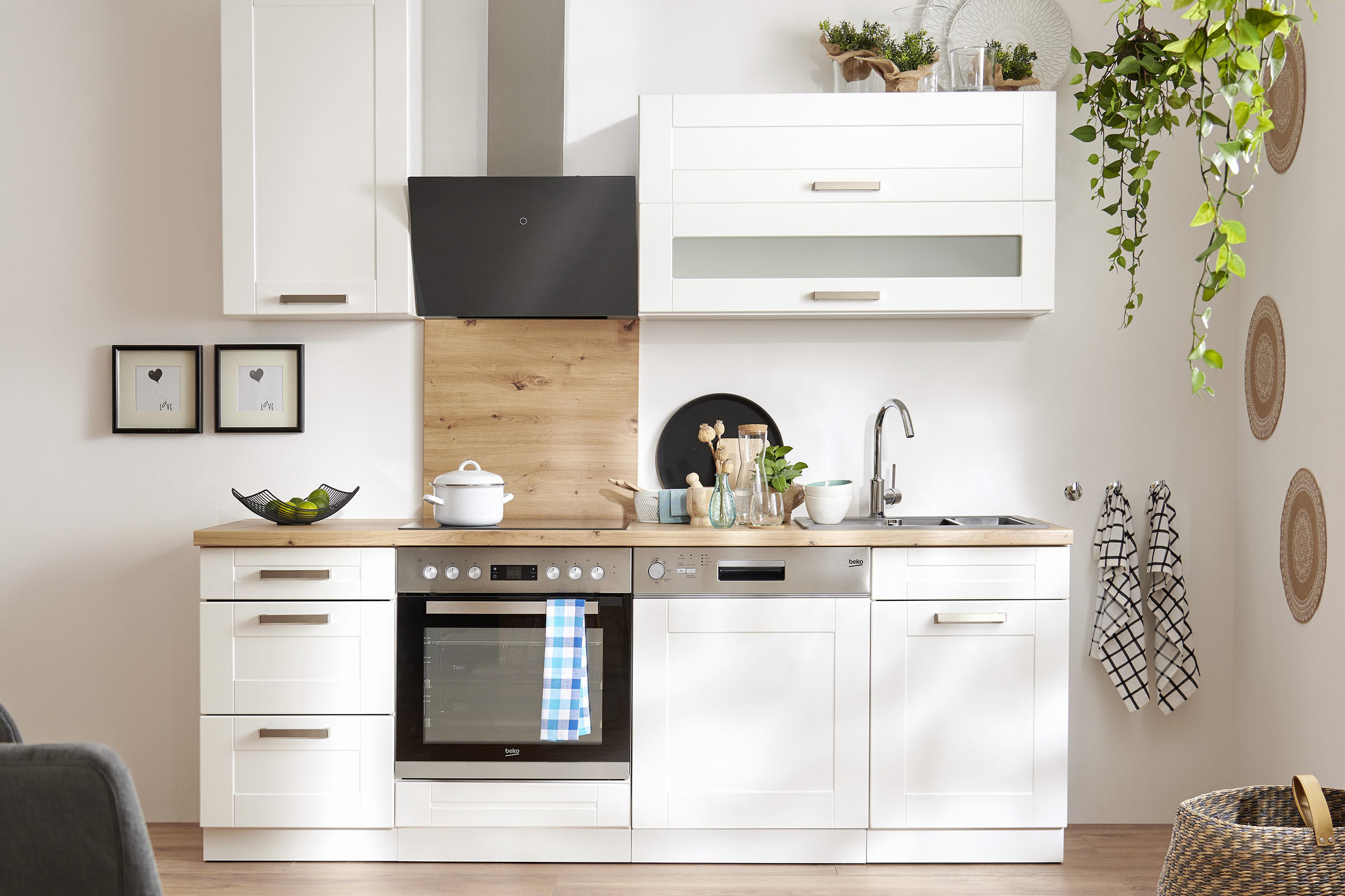 Platzsparende Küchenzeile in Weiß mit Holzarbeitsplatte, schwarzen Dunstabzug und integriertem Backofen; rechts Spüle und Stauraum, dekoriert mit Pflanzen, Geschirr und Handtüchern in einem hellen Wohnraum.
