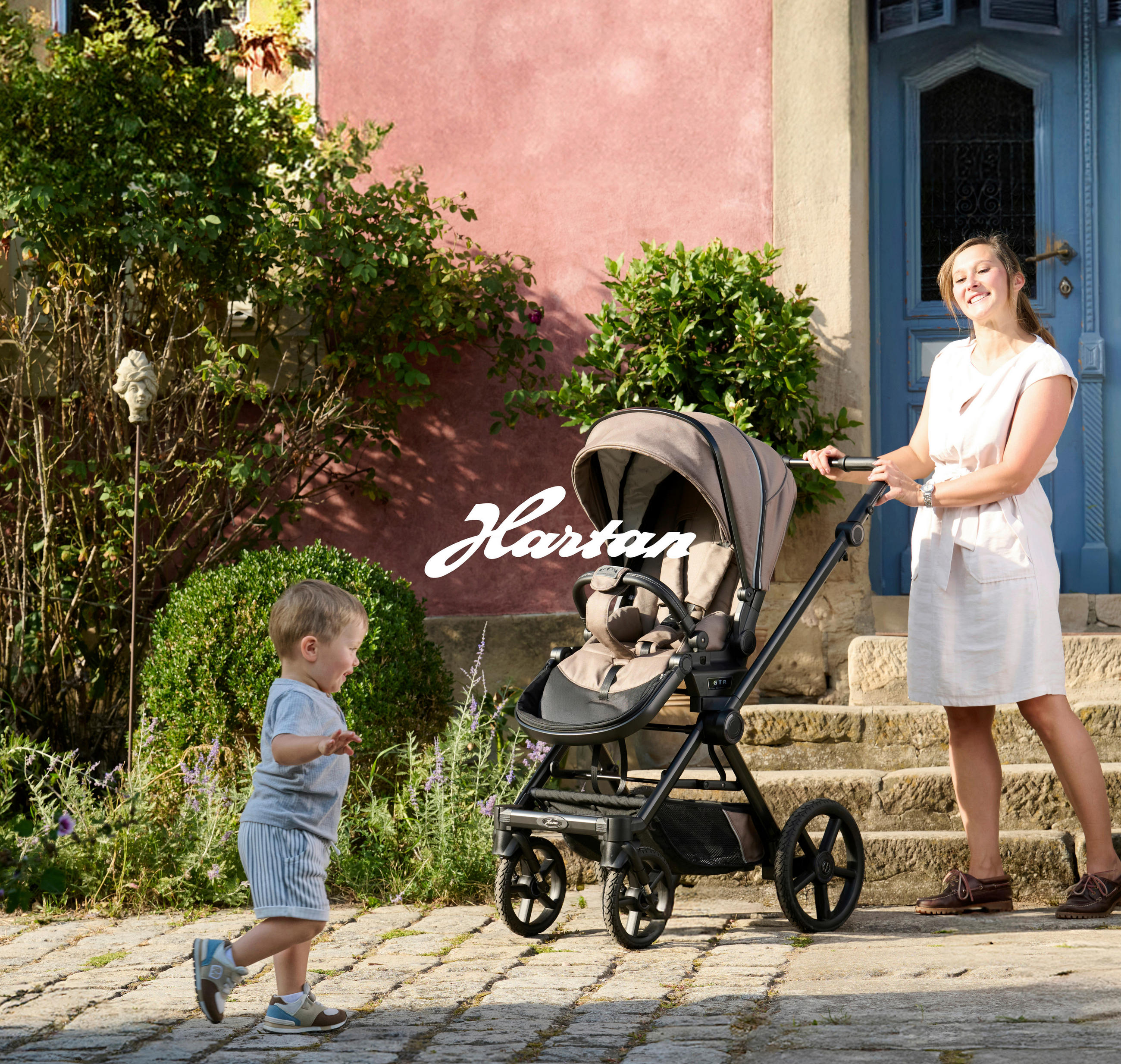 Eine Frau steht vor einem Hauseingang und hält einen Kinderwagen. Im Vordergrund läuft ein kleines Kind über einen sonnigen gepflasterten Weg. Rundherum sind grüne Sträucher und Pflanzen vor einer rosafarbenen Hauswand zu sehen