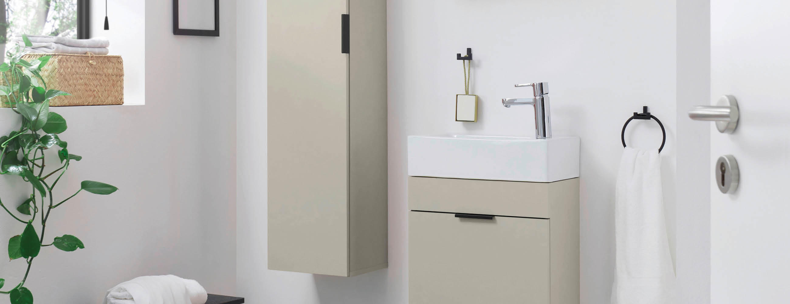 Kleines Badezimmer mit beige-weißem Waschbeckenunterschrank, wandhängendem Spiegelschrank, Handtuchhalter mit weißem Handtuch und grüner Pflanze neben einem Fenster