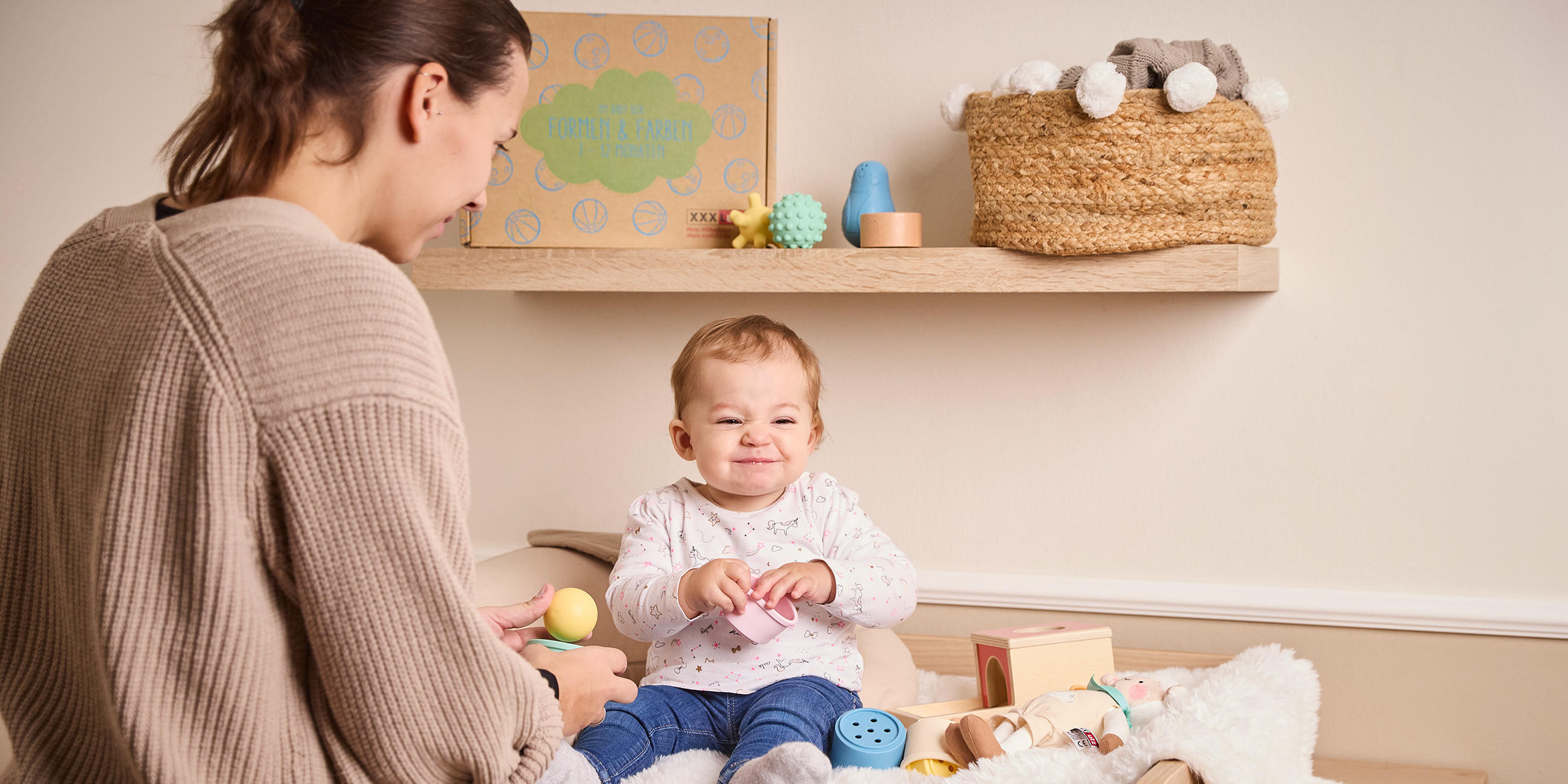 Kind auf Wickelkommode spielt mit Babybox Artikeln während seine Mutter glücklich zusieht