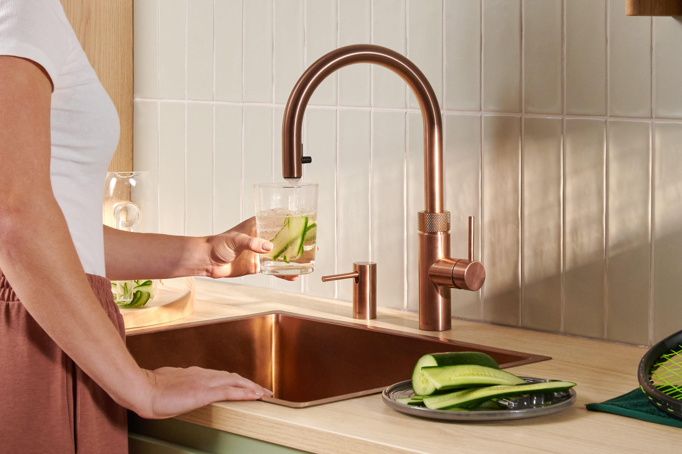 Quooker Wassersystem-Armatur in Kupferoptik über Spülbecken; eine Person füllt ein Glas Wasser mit Gurkenscheiben, daneben liegen frische Gurken auf der Arbeitsfläche.