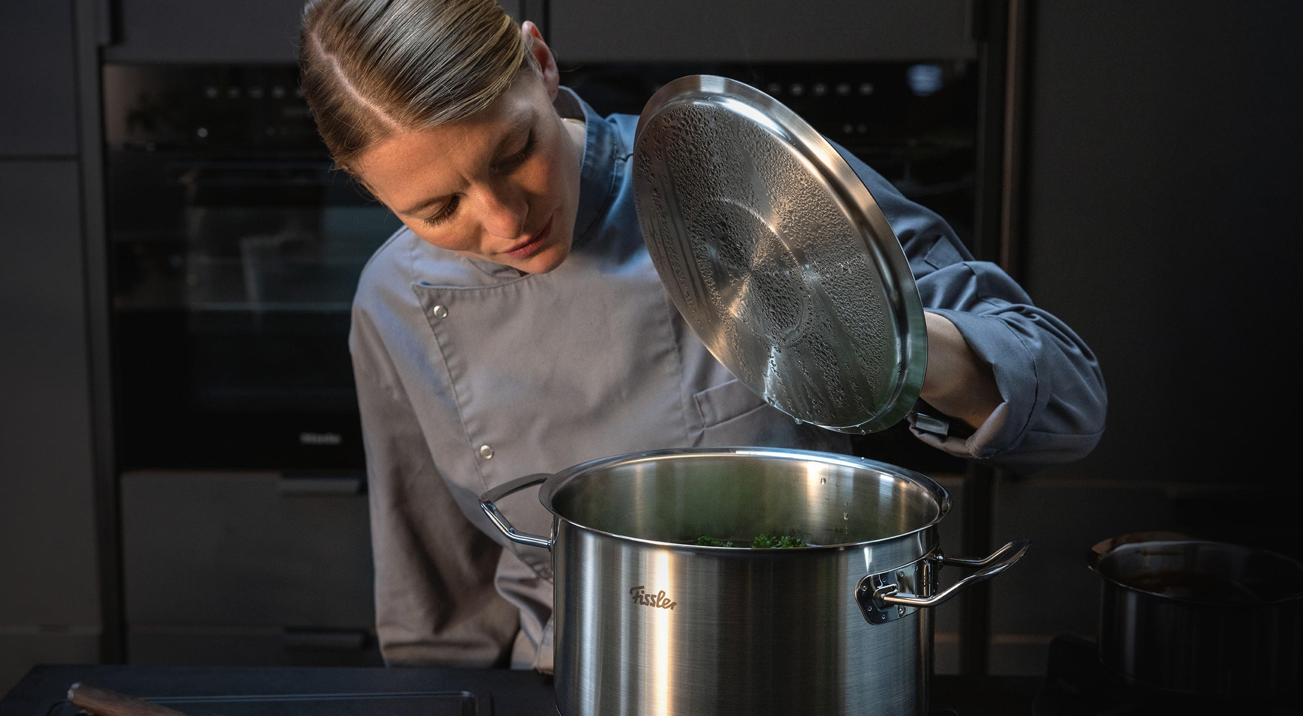 Köchin in grauer Kochjacke hebt den beschlagenen Deckel eines großen Edelstahl‑Topfs an; auf dem Topf ist das FISSLER‑Logo sichtbar, im dunklen Küchenhintergrund stehen weitere Töpfe.