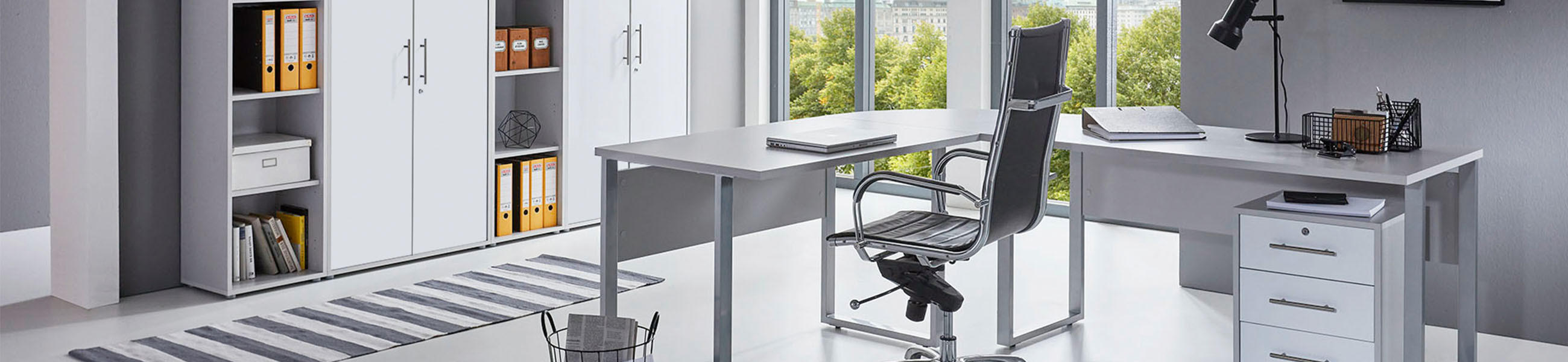 Modernes Büro mit weißem Eckschreibtisch, schwarzem Bürostuhl, Rollcontainer und weiß matten Aktenschränken vor großen Fenstern.