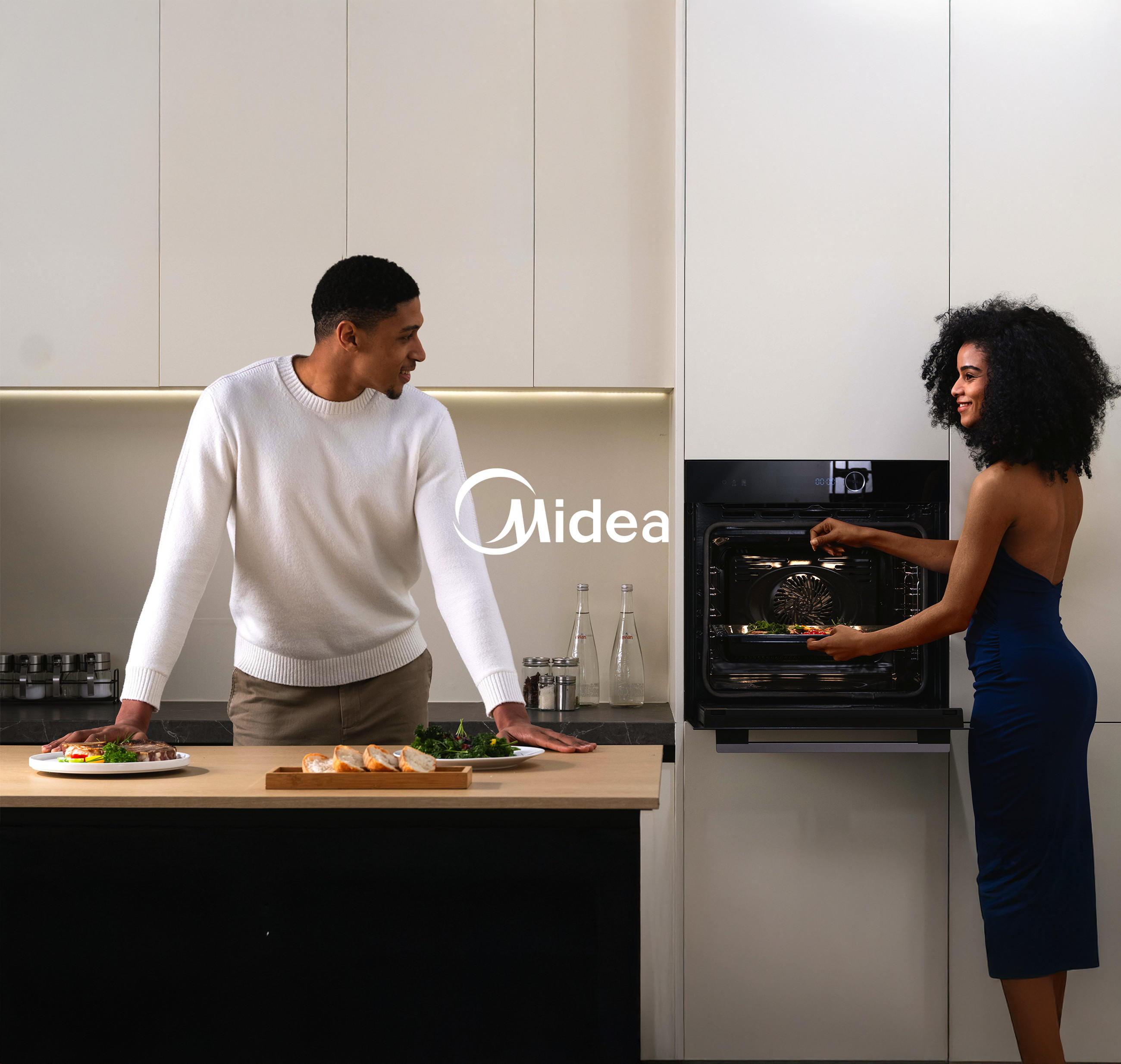 Moderne Küche: Frau schiebt ein Backblech in einen eingebauten Midea Backofen, Mann schaut lächelnd zu; zentral eingeblendetes Midea-Logo.