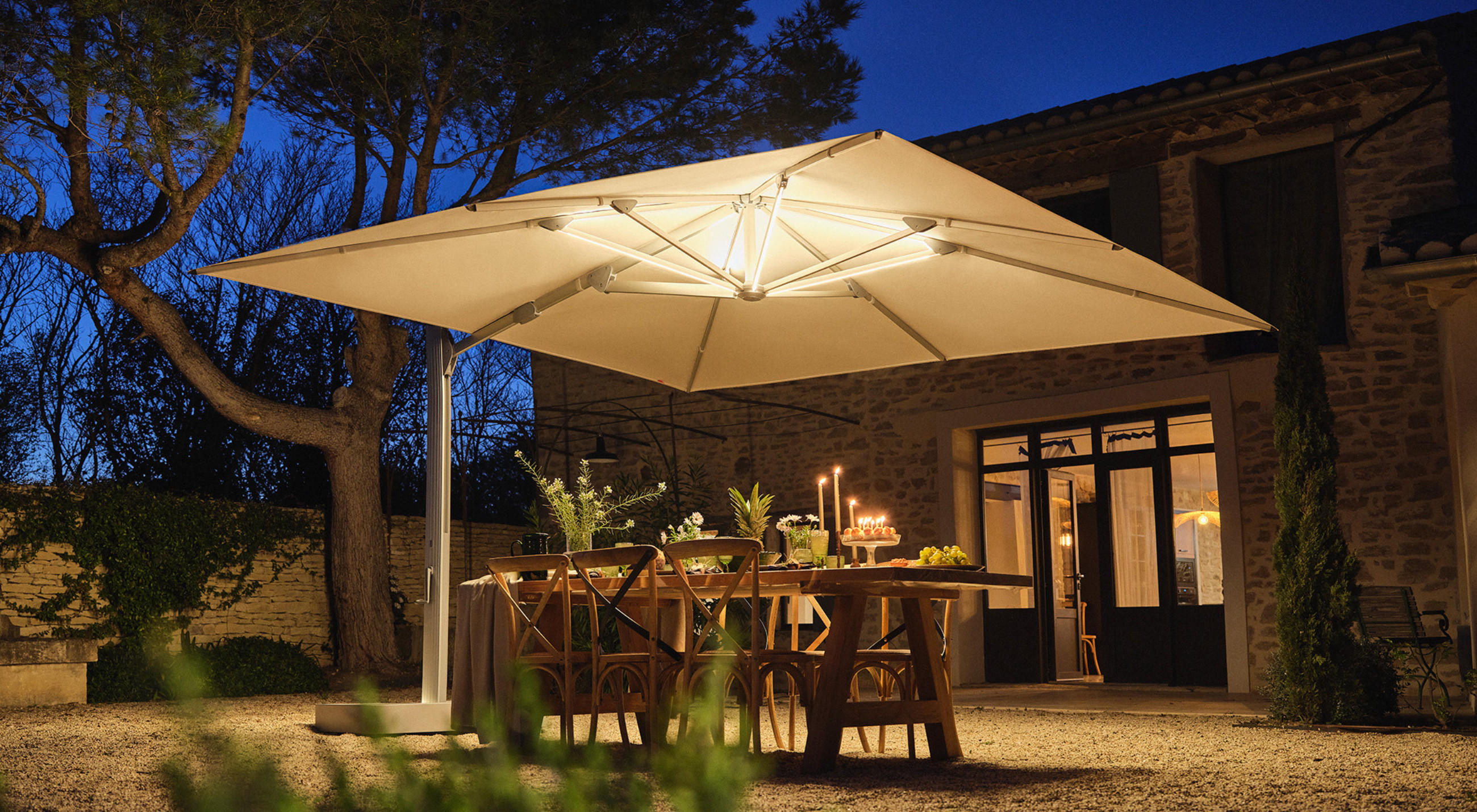 Ein großer Terrassenschirmvon GLATZ mit eingebauter Beleuchtung spendet Schatten über einem Esstisch im Freien, der für ein Abendessen in einem Innenhof in der Abenddämmerung gedeckt ist.