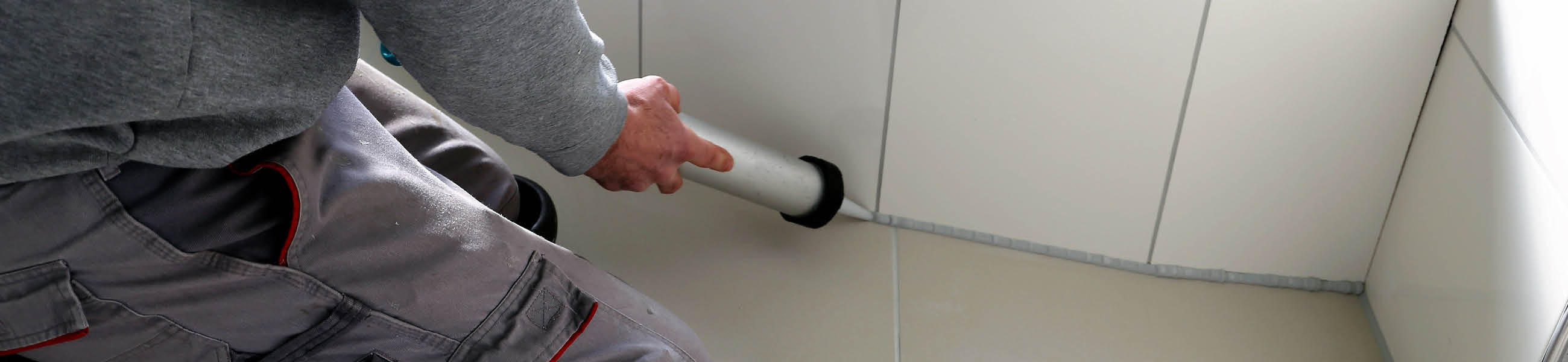 Person in grauem Sweatshirt und Arbeitshose, die eine Dichtungsmasse in einen Wandspalt aufträgt, um eine Fliese zu reparieren.