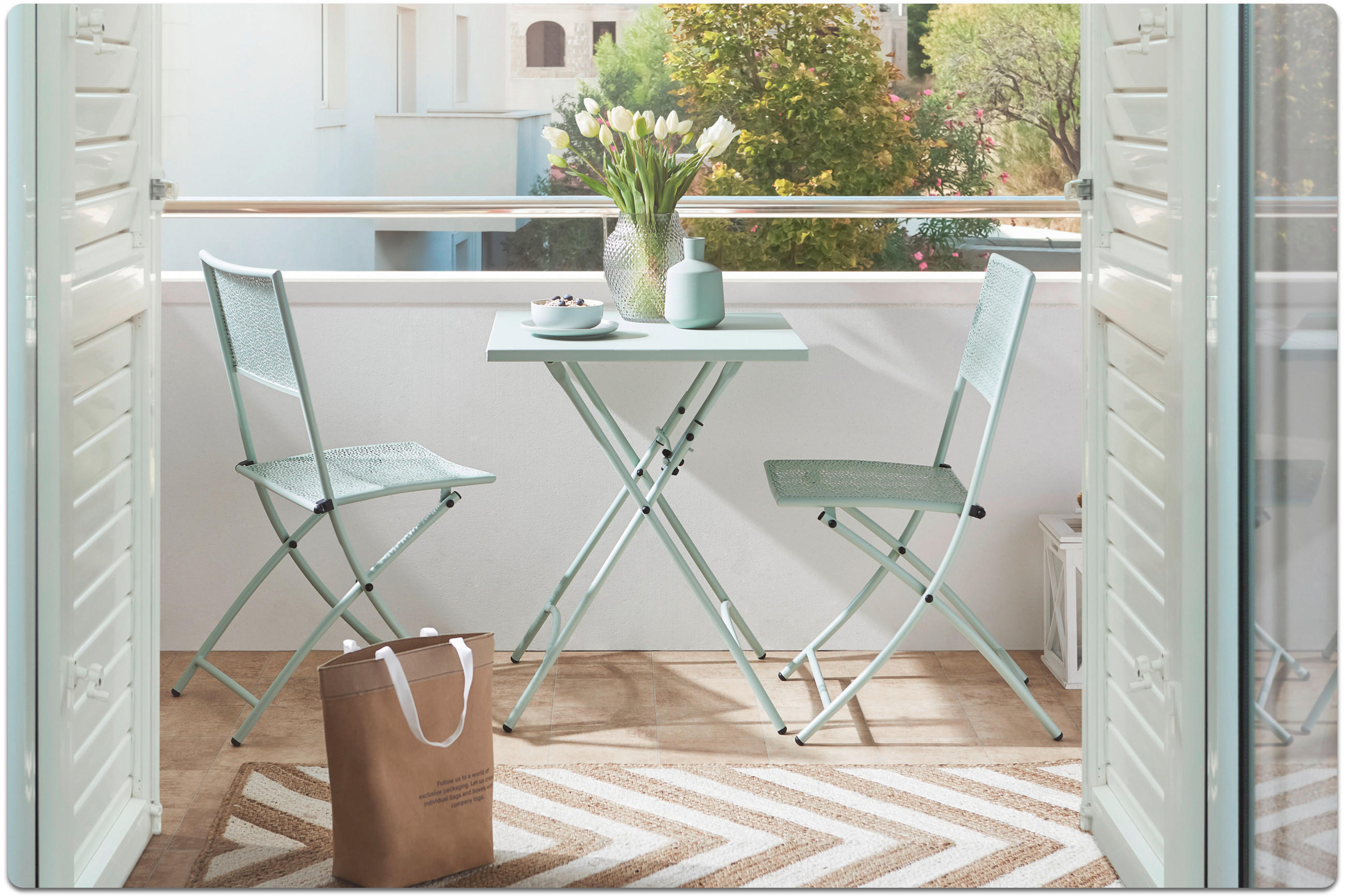 Balkon mit kleinem weißen Klapptisch, zwei weißen Klappstühlen, Vase mit weißen Tulpen und brauner Papiertüte auf dem Boden.
