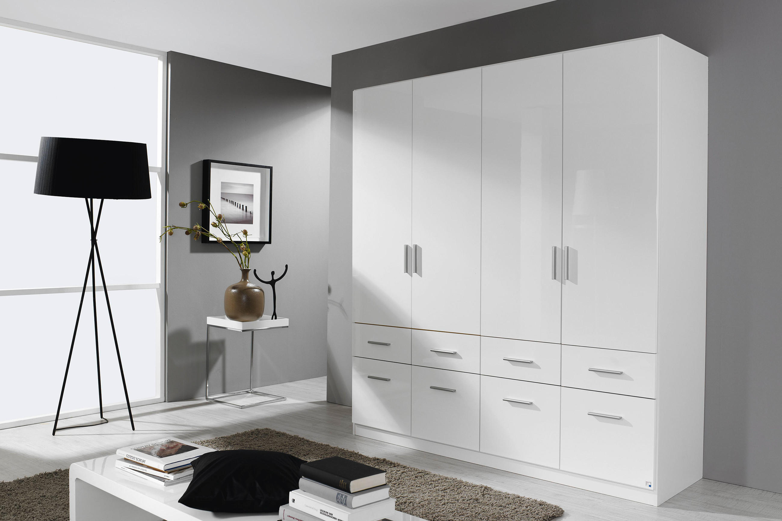 Weißer Kleiderschrank mit Türen und Schubladen in modernem, hellem Raum; daneben schwarze Stehlampe, Beistelltisch mit Vase und Bild.