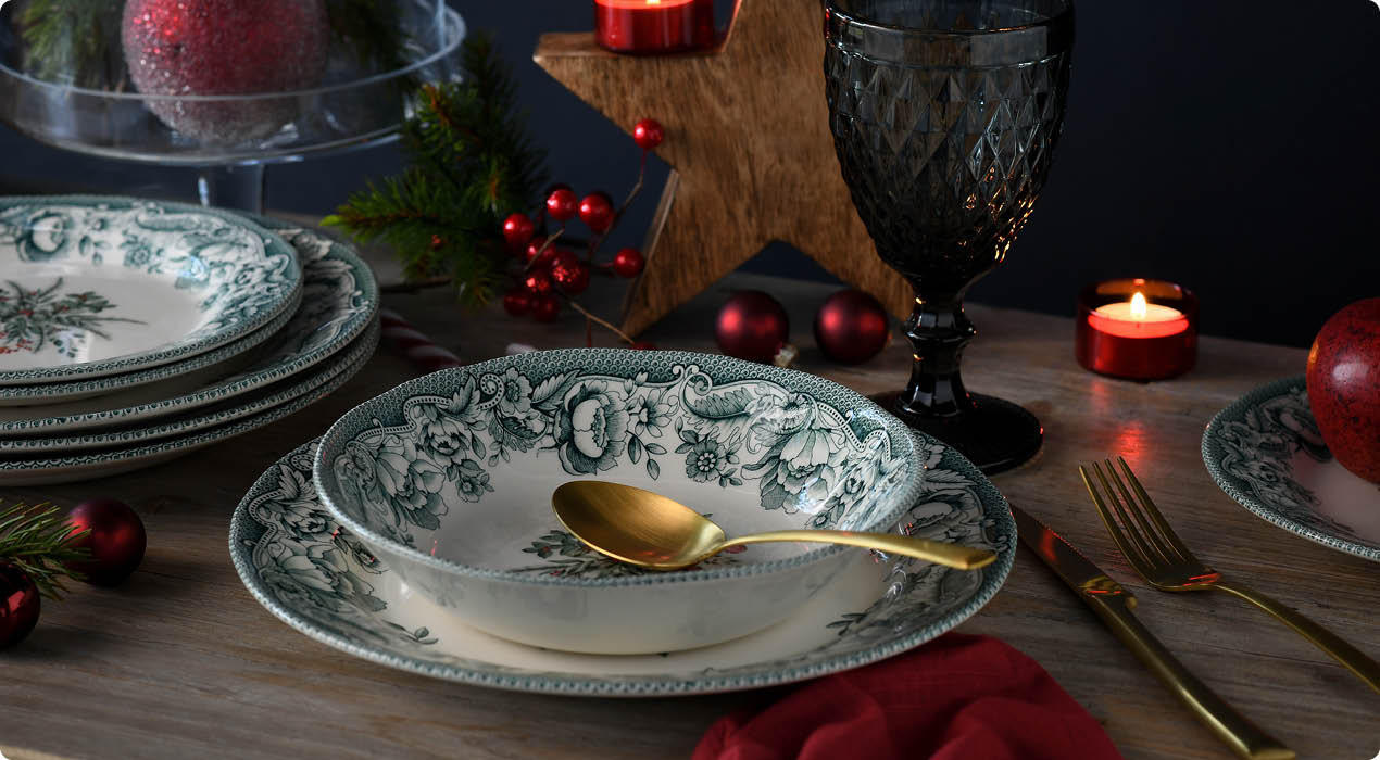 Esstisch mit grünen, floral gemusterten Porzellantellern und -schalen, goldfarbenem Besteck, roten Weihnachtskugeln, Kerzen und einem roten Stoffserviette als Dekoration.