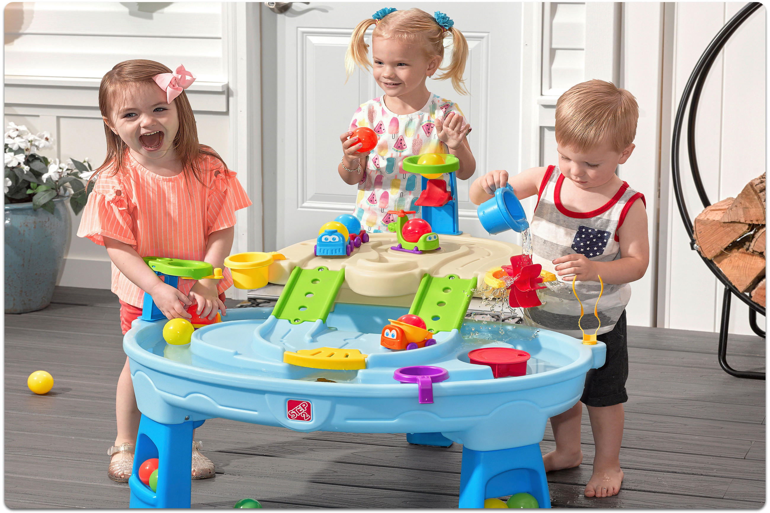 Drei Kinder spielen an einem blauen Wasserspieltisch mit buntem Zubehör auf einem Balkon.