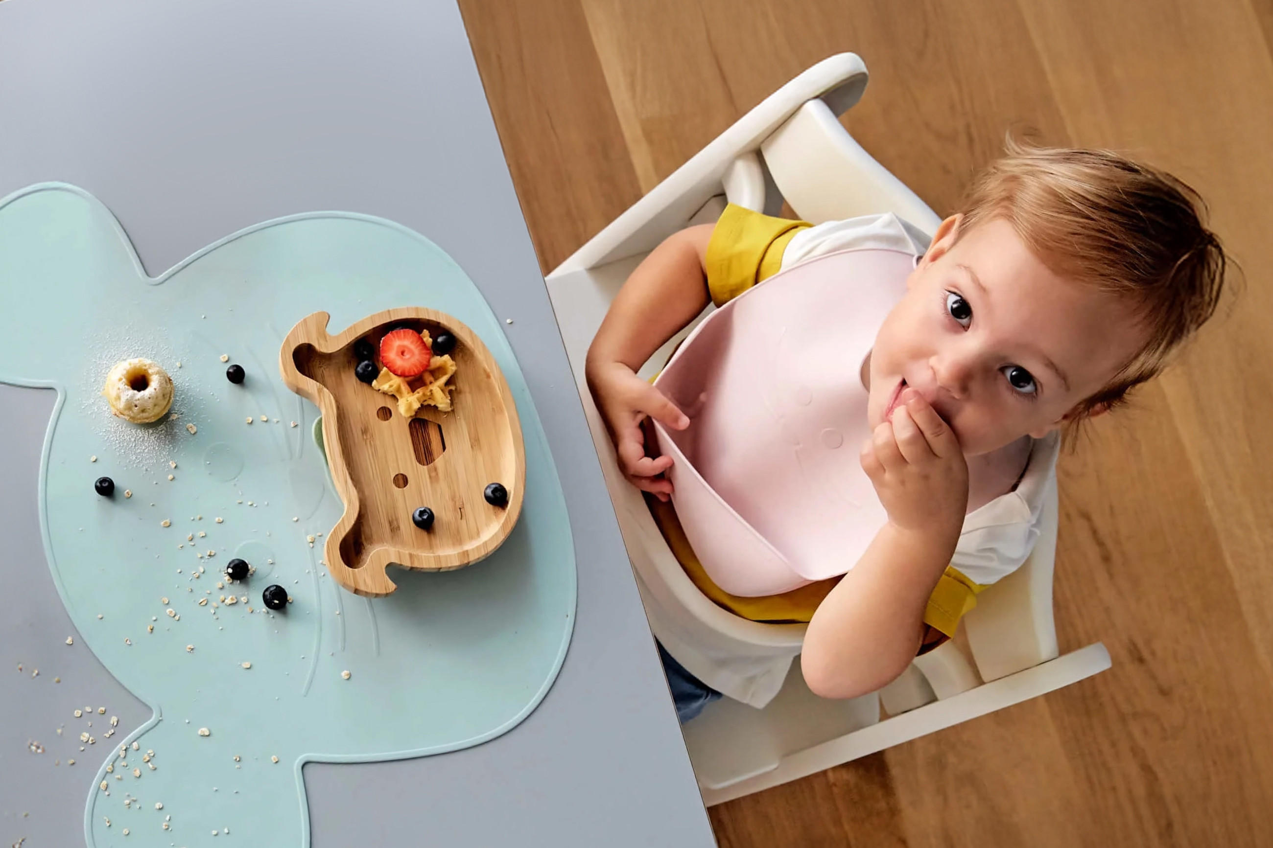 Kind sitzt auf einem Hochstuhl am Tisch mit einem wolkenförmigen Platzset und einem Holztablett in Tierform mit etwas Essen.