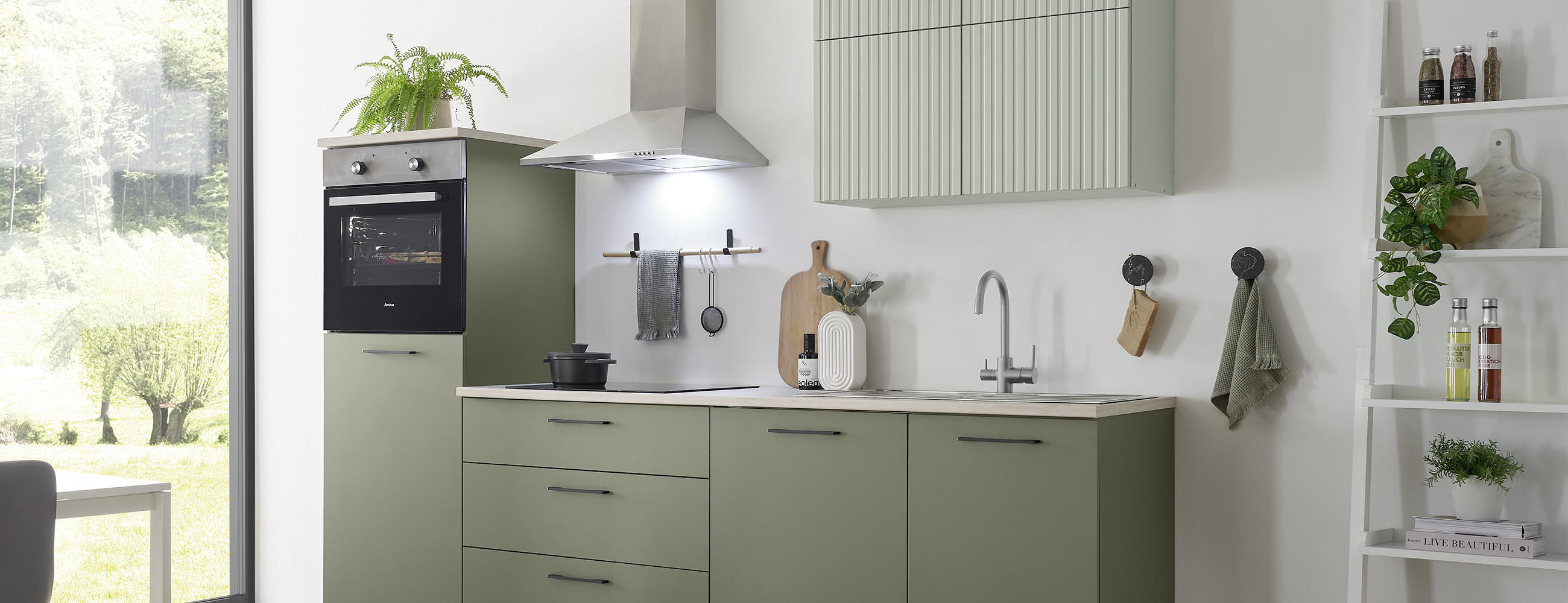 Helle, kleine Küche mit grünen Fronten und Holzarbeitsplatte, integriertem Backofen im Hochschrank sowie Dunstabzug über dem Kochfeld; daneben Spüle, offene Regale mit Flaschen und Pflanzen und großes Fenster mit Blick ins Grüne.