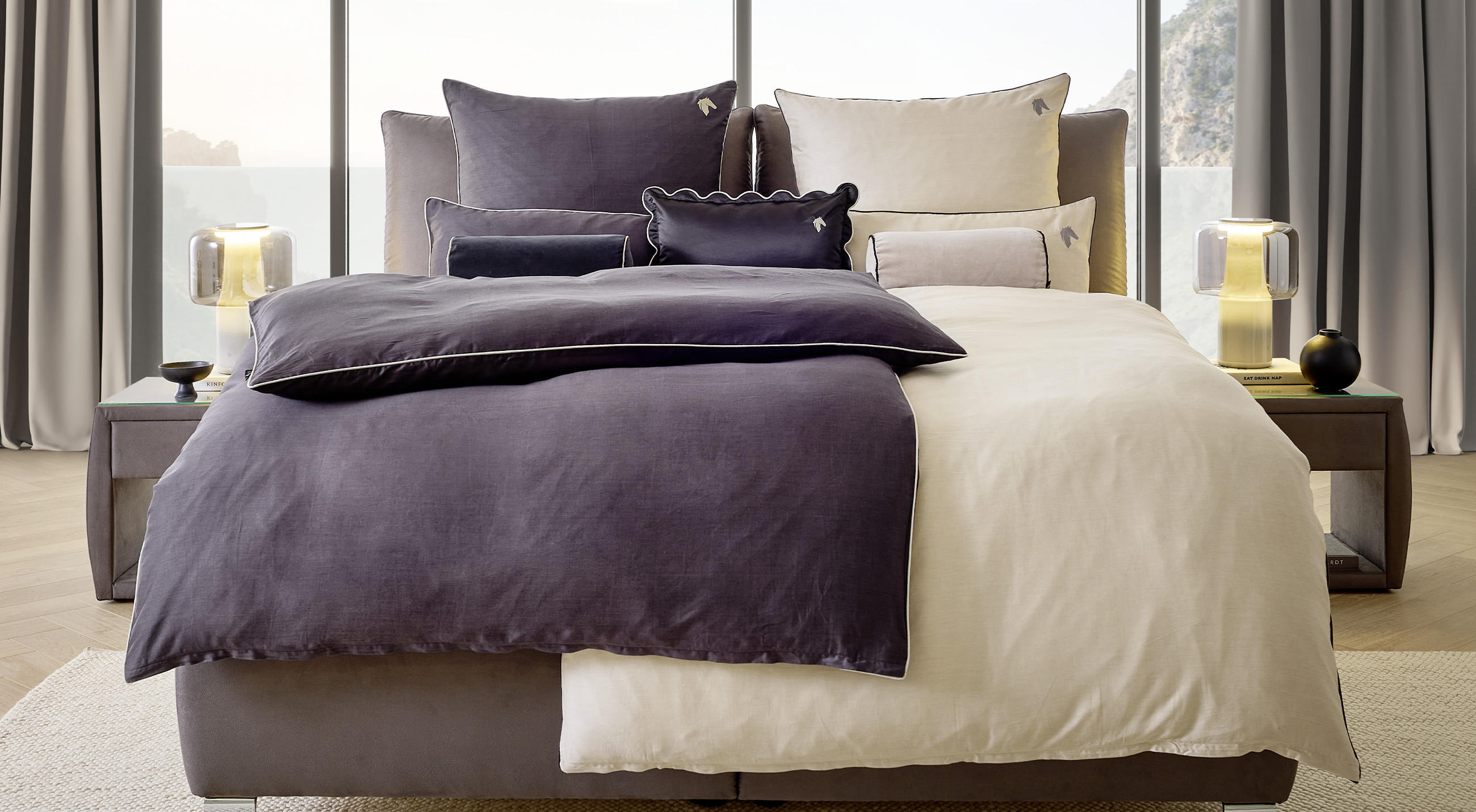 Modernes Schlafzimmer mit großem Bett in Beige- und Dunkelgrautönen, dekoriert mit mehreren Kissen und einer Tagesdecke. Rechts und links Nachttische mit Lampen und Deko, im Hintergrund große Fenster mit Berg- und Meerblick