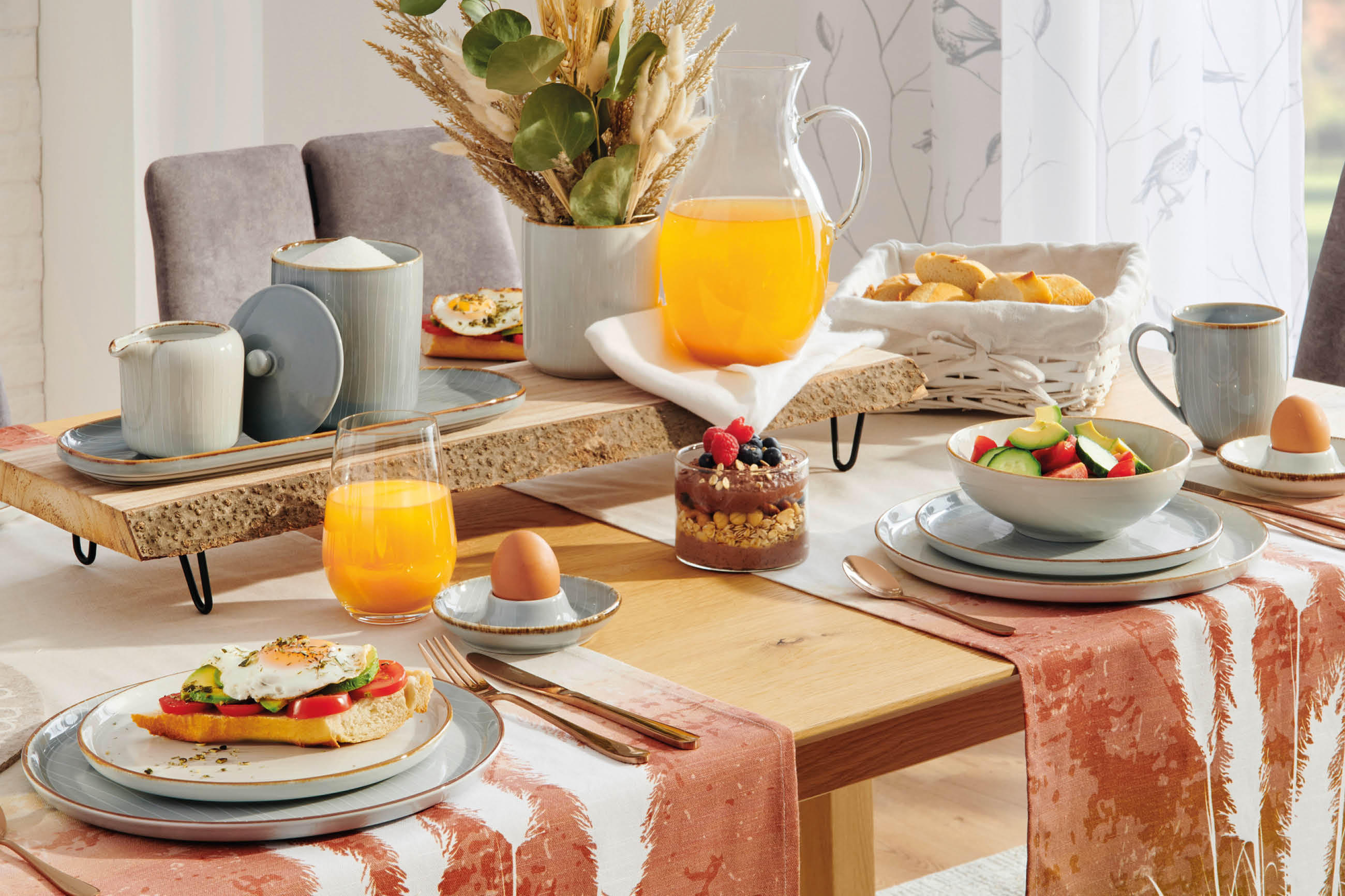 Esstisch mit gedeckten Tellern, Gläsern mit Orangensaft, Eiern in Eierbechern, einem Dessertglas, Brotkorb und einer Vase mit Trockenblumen auf einem Tischläufer