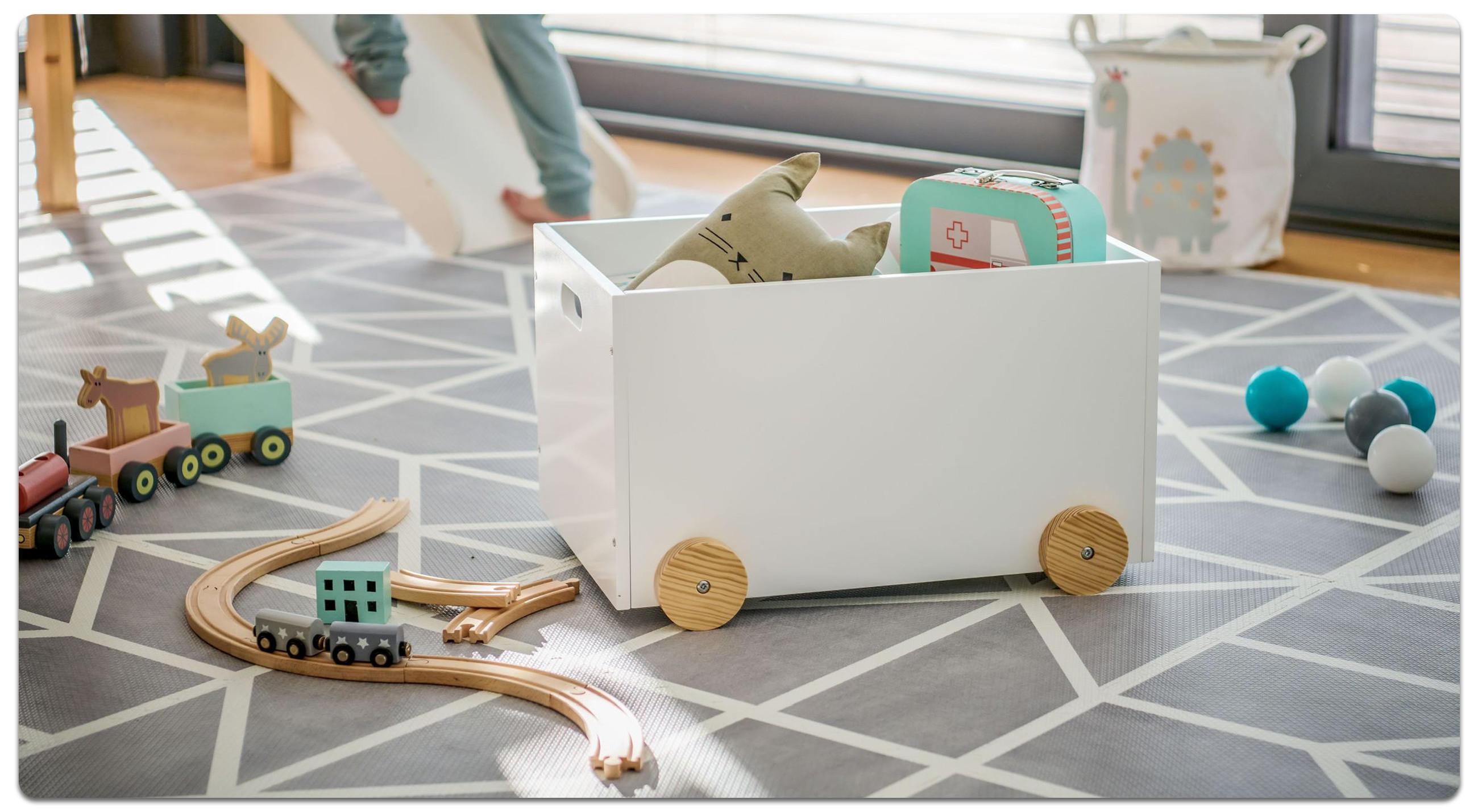 Eine gefüllte Spielzeigbox mit Rollen neben Holzfahrzeugen auf Schienen und ein Kind benutzt eine Rutsche im Hintergrund.