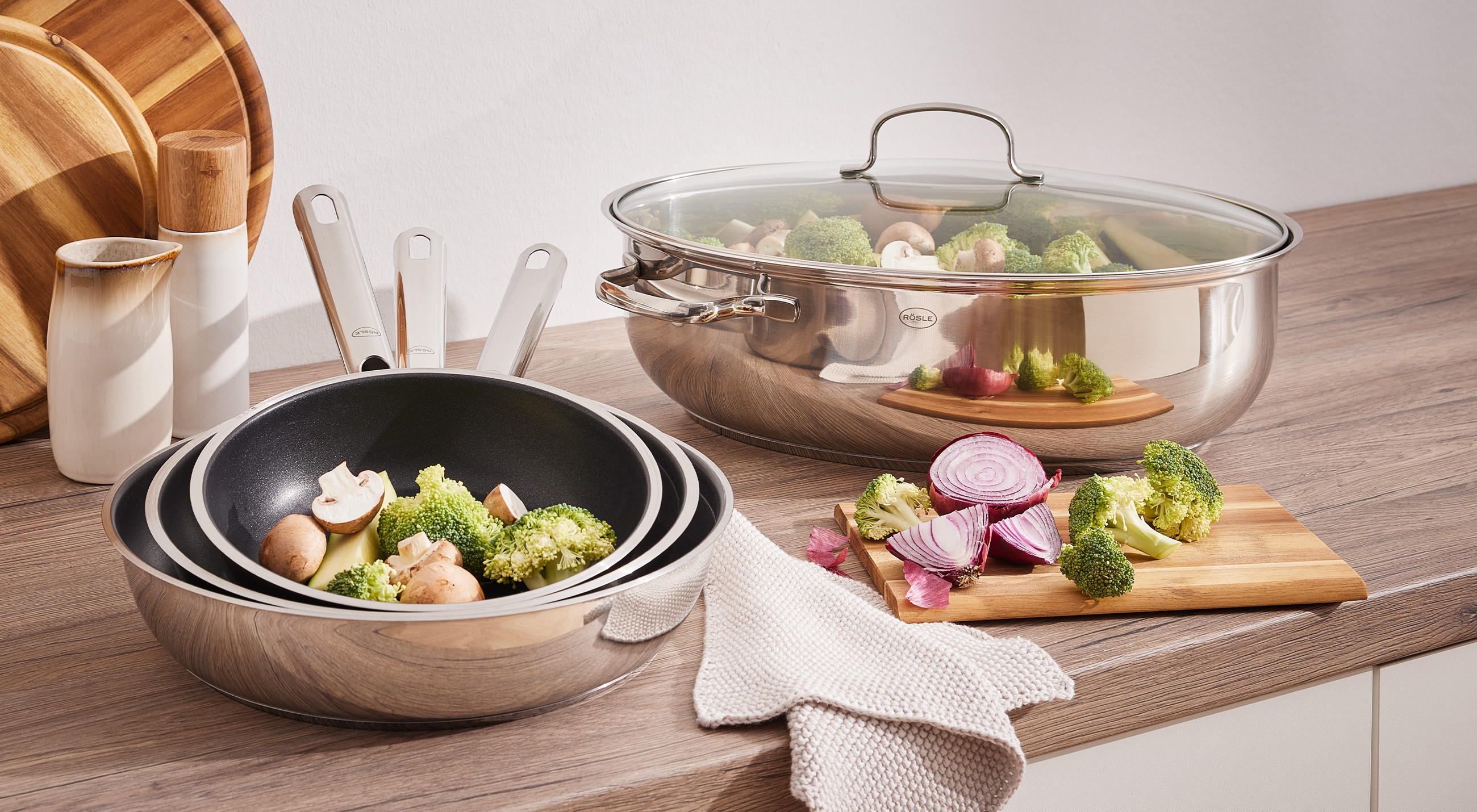 Küchenplatte mit Edelstahl‑Kochgeschirr: großer Topf mit Glasdeckel mit Brokkoli und Champignons, drei gestapelte Antihaft‑Pfannen, Holz‑Schneidebrett mit halbierter roter Zwiebel, Keramikbehälter und Holz‑Schneidebretter im Hintergrund